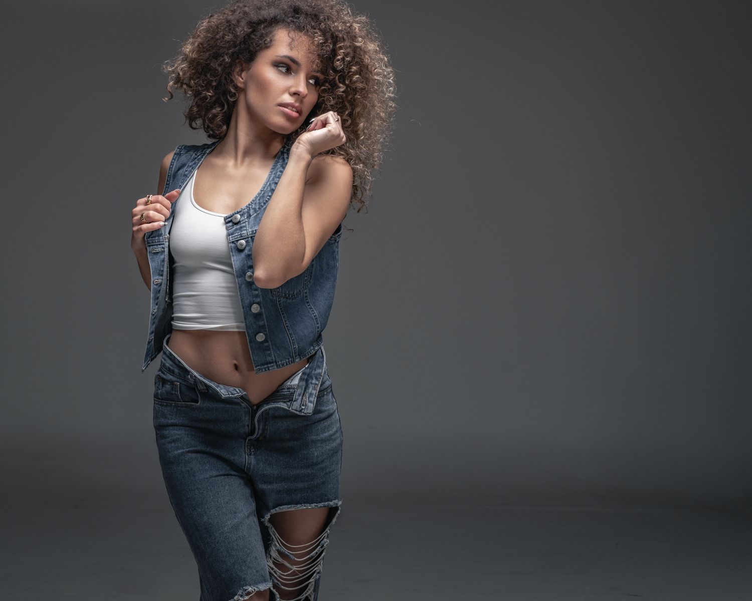 Young woman with curly hair posing in ripped jeans, denim vest, and white crop top against gray background in the studio.