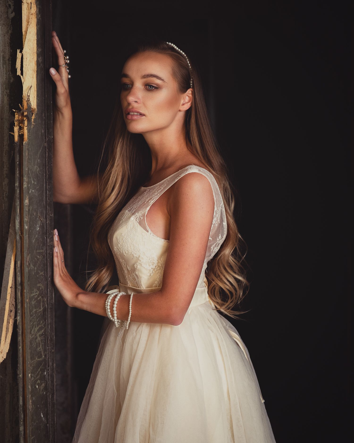 Global fashion photographer portfolio: detailed cream dress with pearls, model with long wavy hair leans on rustic wooden door frame.