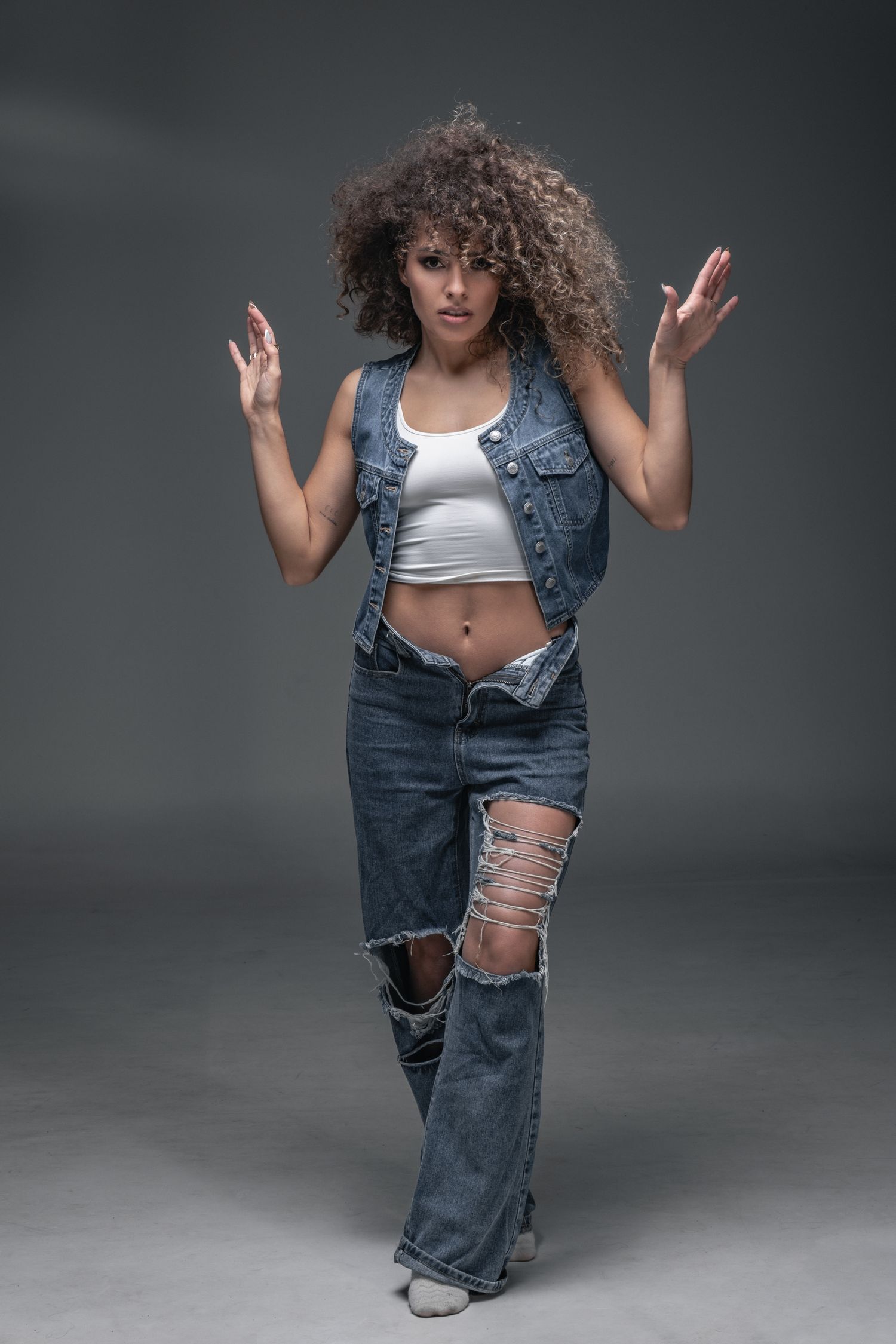 Woman with curly hair posing in ripped jeans, denim vest, and white crop top against gray background.