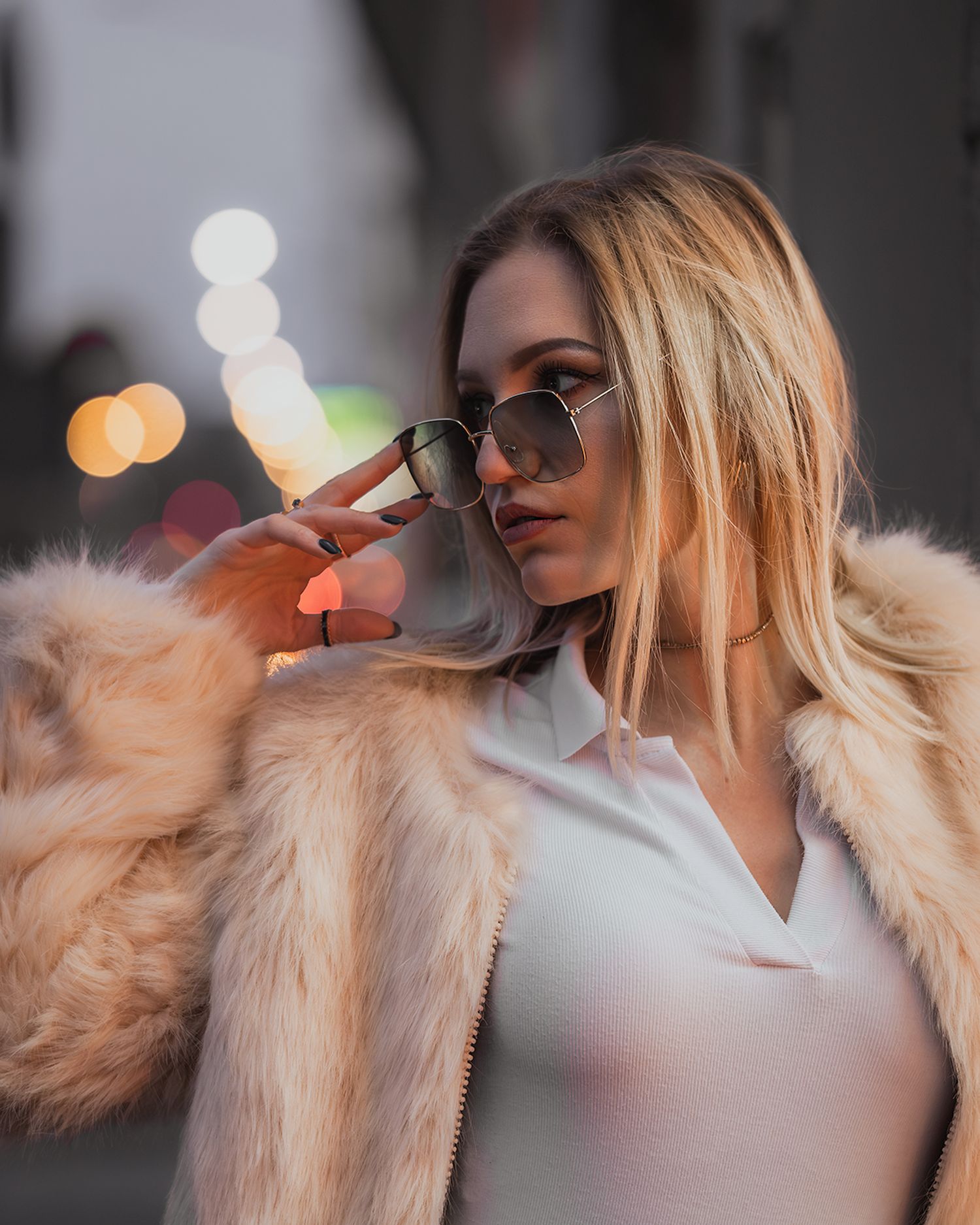 Professional street style photography: model in pink fur jacket looking away, city lights at dusk background, artistic bokeh.