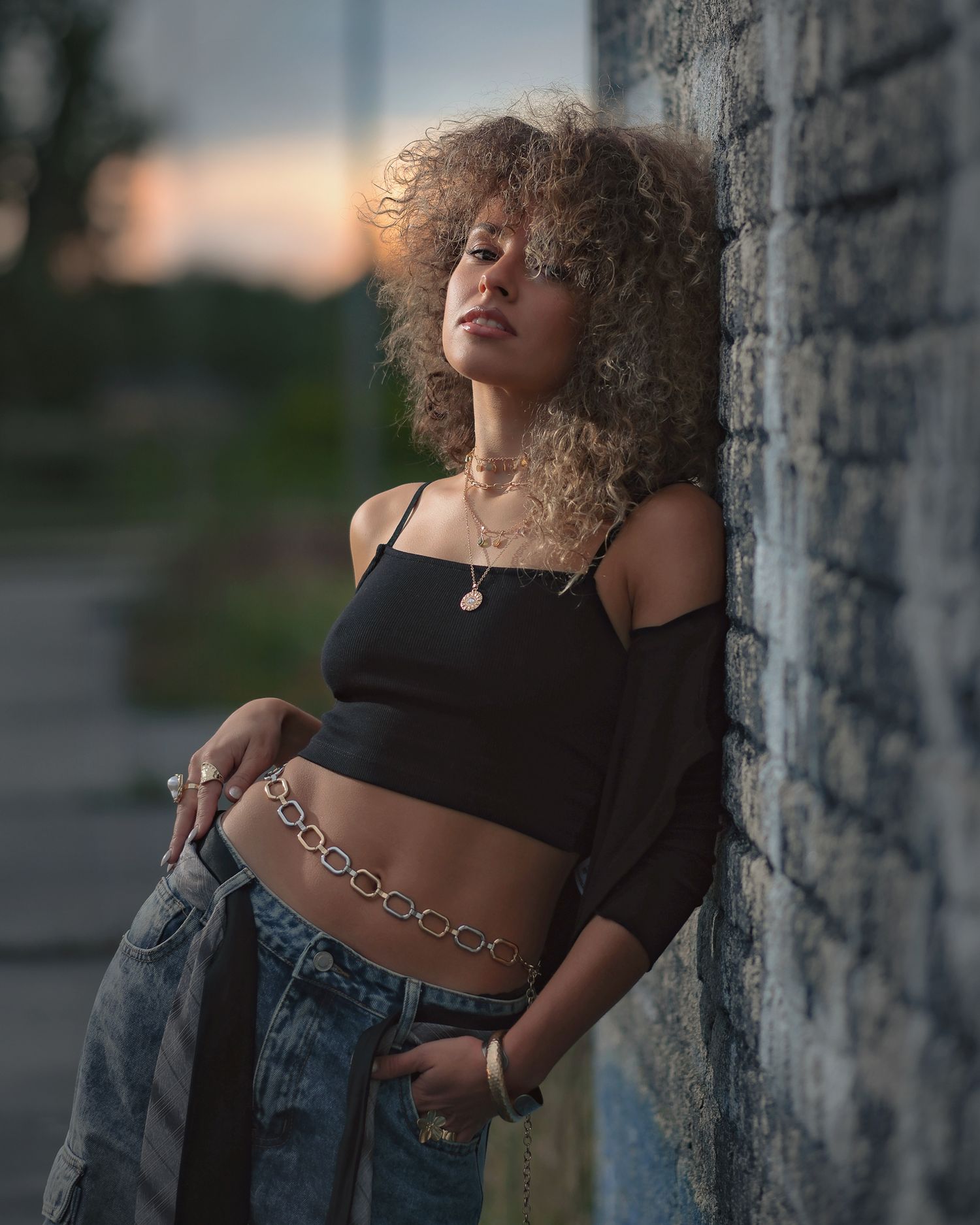Professional street style shot: model in a black crop top and denim, leaning on a dark textured wall, sunset bokeh in background.