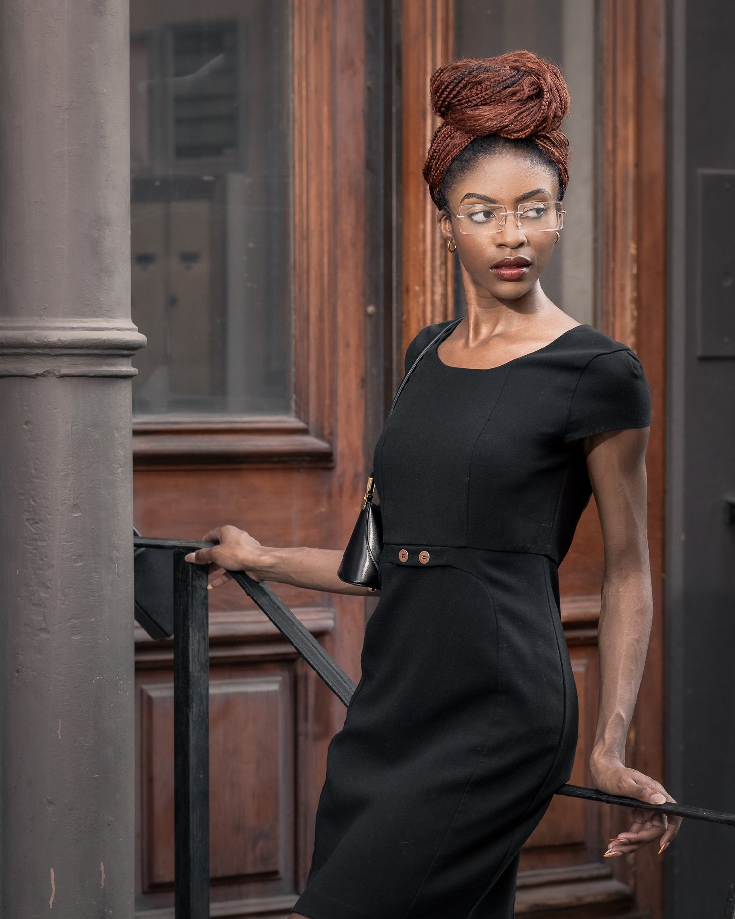 Professional fashion portrait, mid-length crop: model with long copper braids, rimless glasses, sleek black dress and small purse.