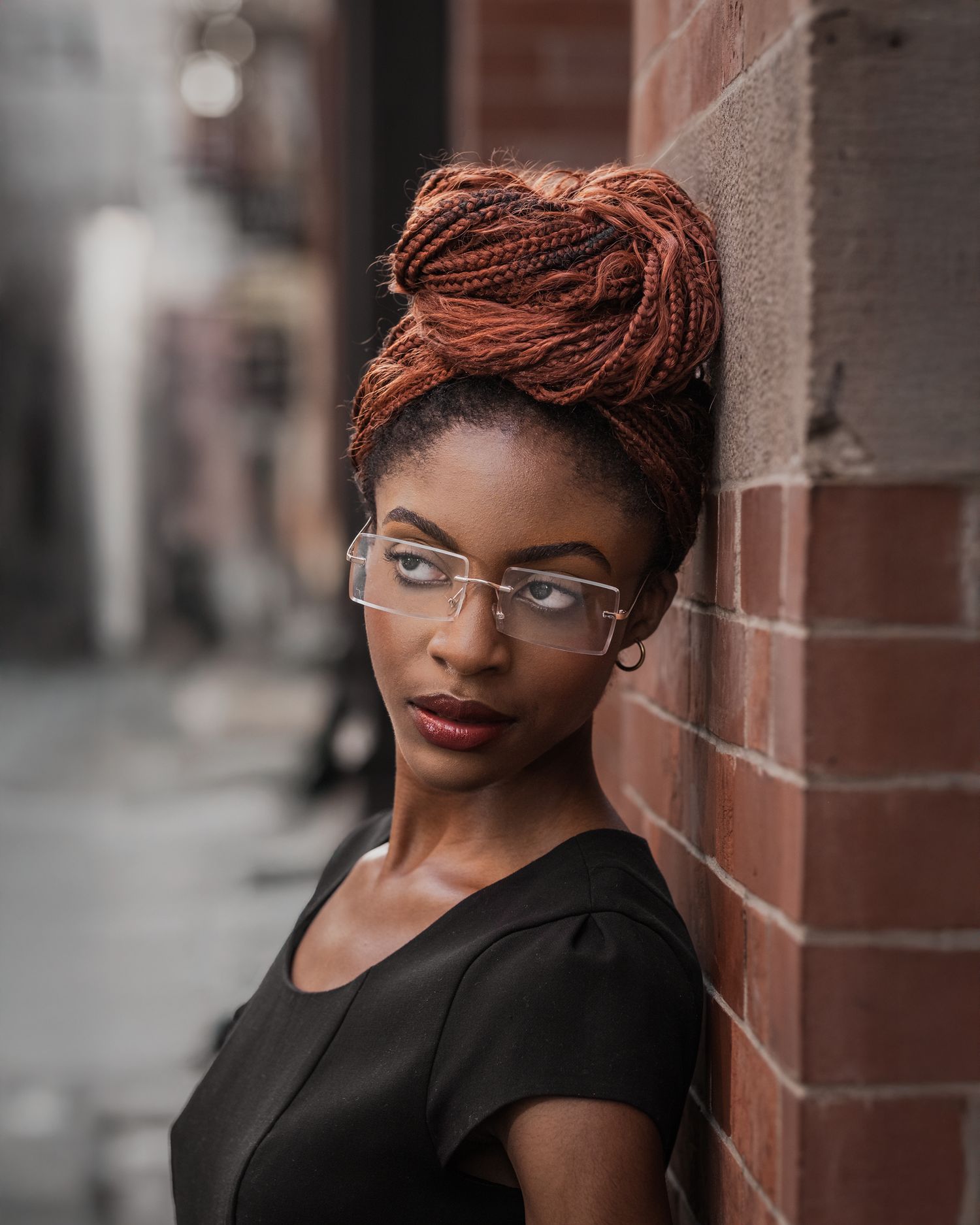Editorial headshot: model with long red braids and clear glasses looking away, serious expression, brick background texture.