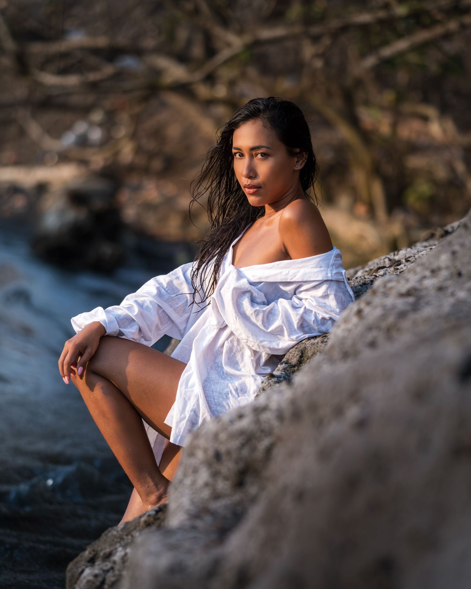 Woman in white off-shoulder shirt sitting on rocks near a stream in a natural outdoor setting.