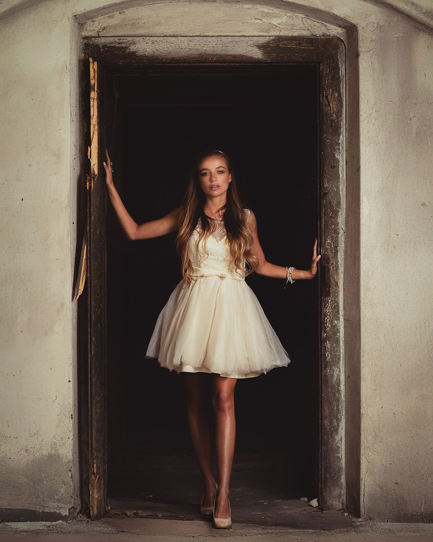 Fashion portrait: model in cream lace & tulle mini dress posing in dark weathered doorway, high-contrast, moody atmosphere.