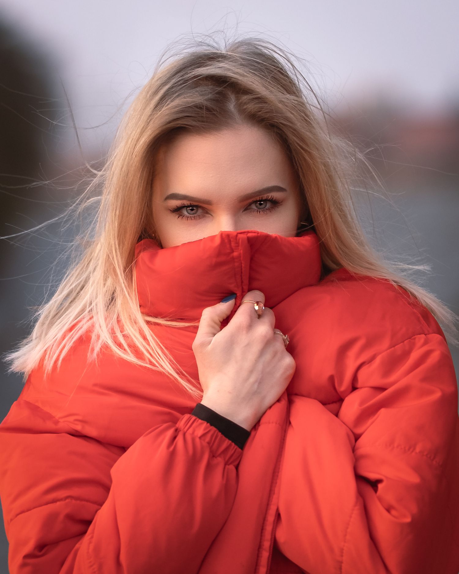 Blonde woman in red puffer jacket covering face, gazing at camera outdoors.