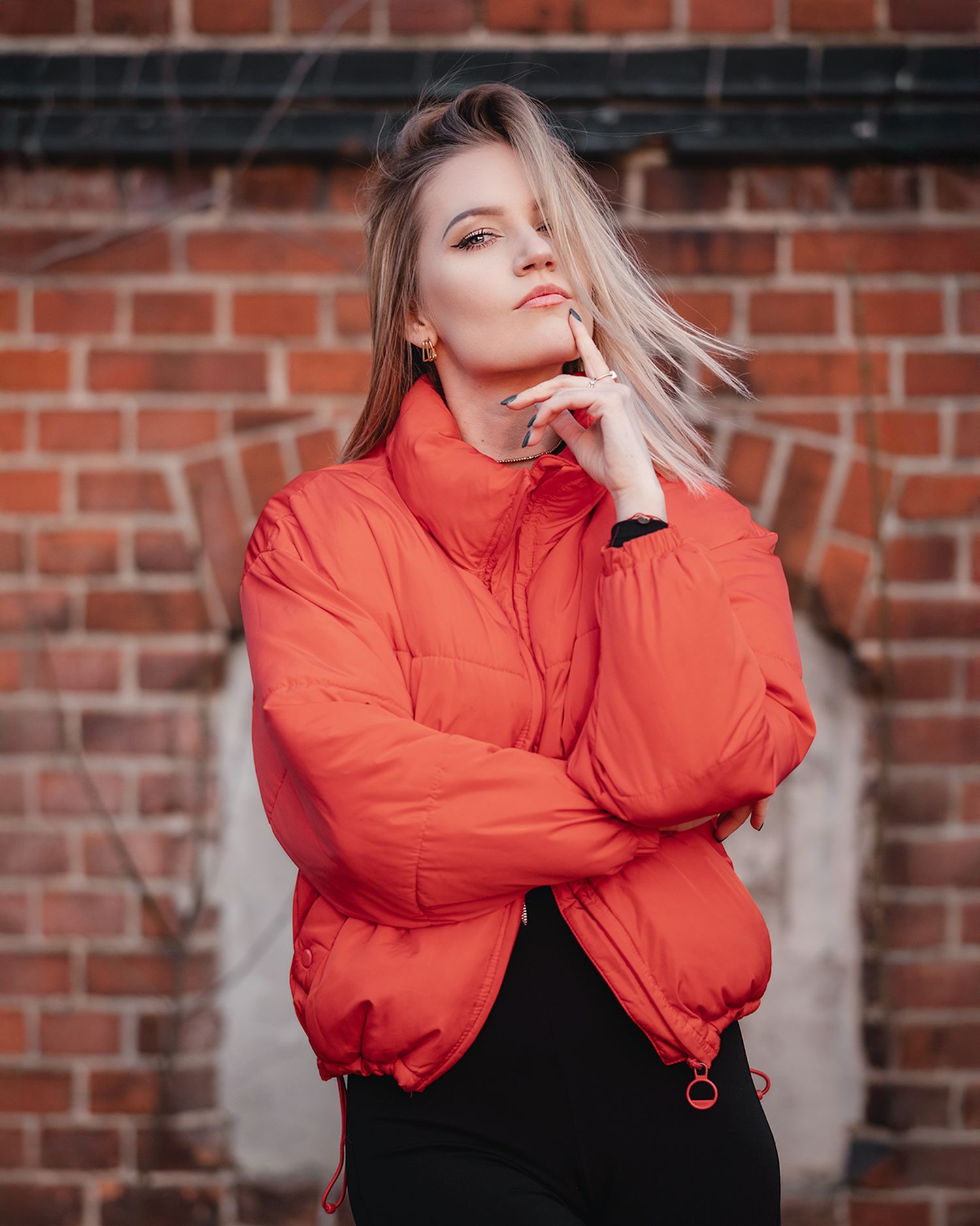 Young blonde woman in a bold red puffer jacket poses thoughtfully against a rustic brick wall.