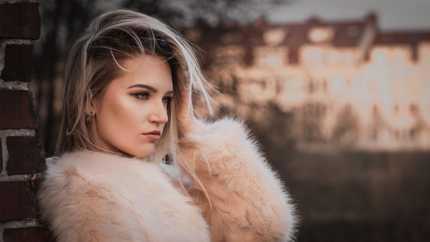 Cinematic urban fashion: model with wind-blown hair in pink fur coat, touching hair, historic building background at sunset.