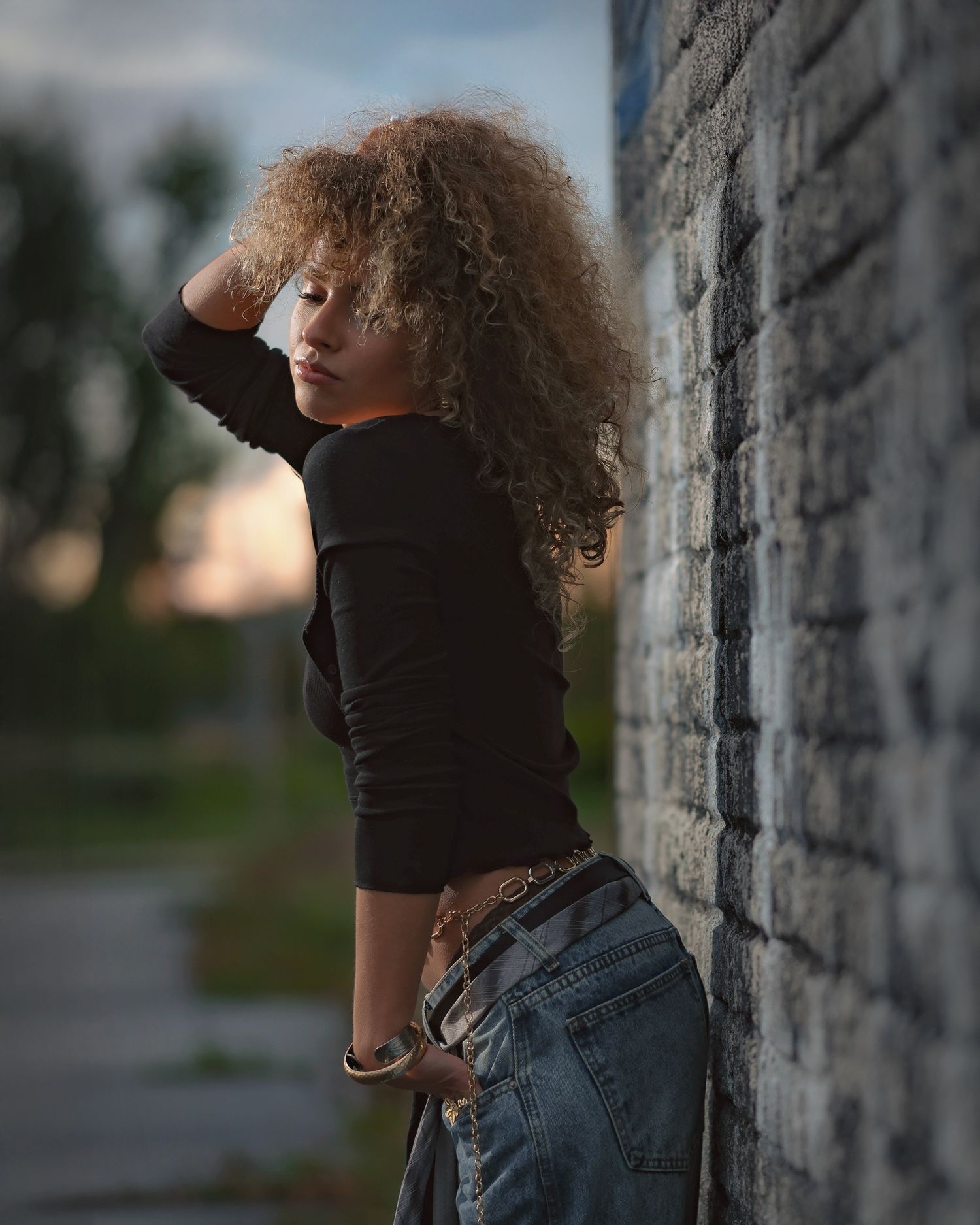Moody urban lifestyle fashion: profile of a model with large curly hair, detailed gold rings and bracelets, hand touching face.