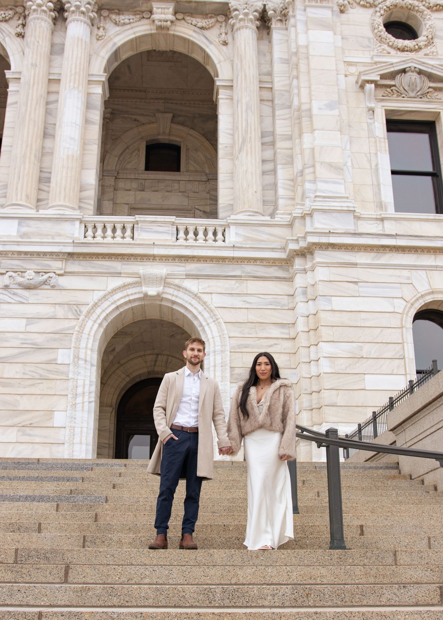 A couple stands on the grand stone steps of an ornate classical building with arched windows and architectural details.