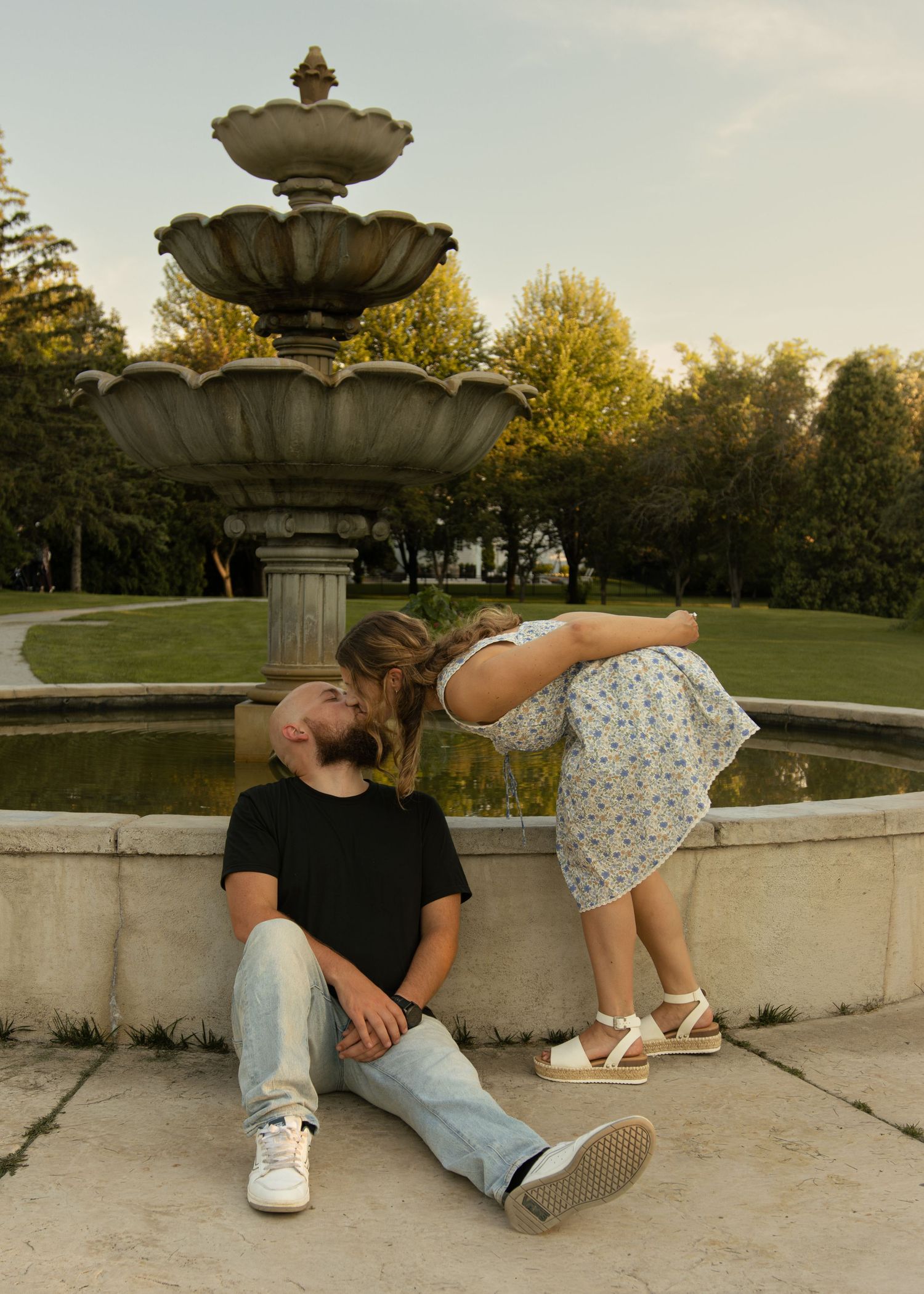 Couple sharing a kiss by a stone fountain in a park, man sitting on ground in black tee and jeans.