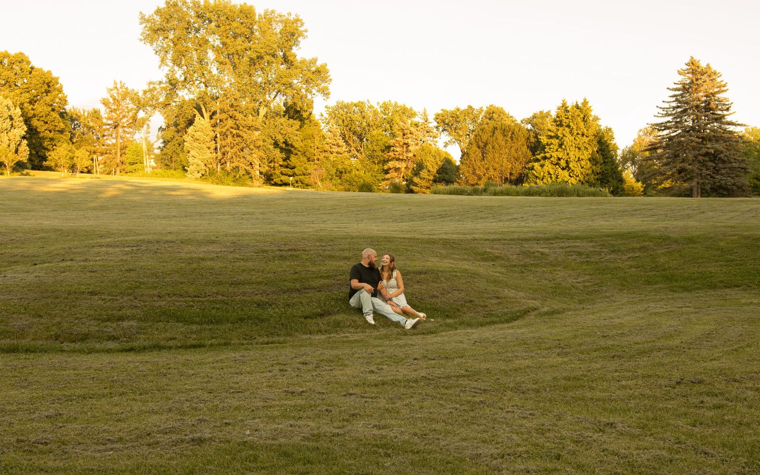 Couple sitting together on a grassy hillside at golden hour with lush green trees in the background.