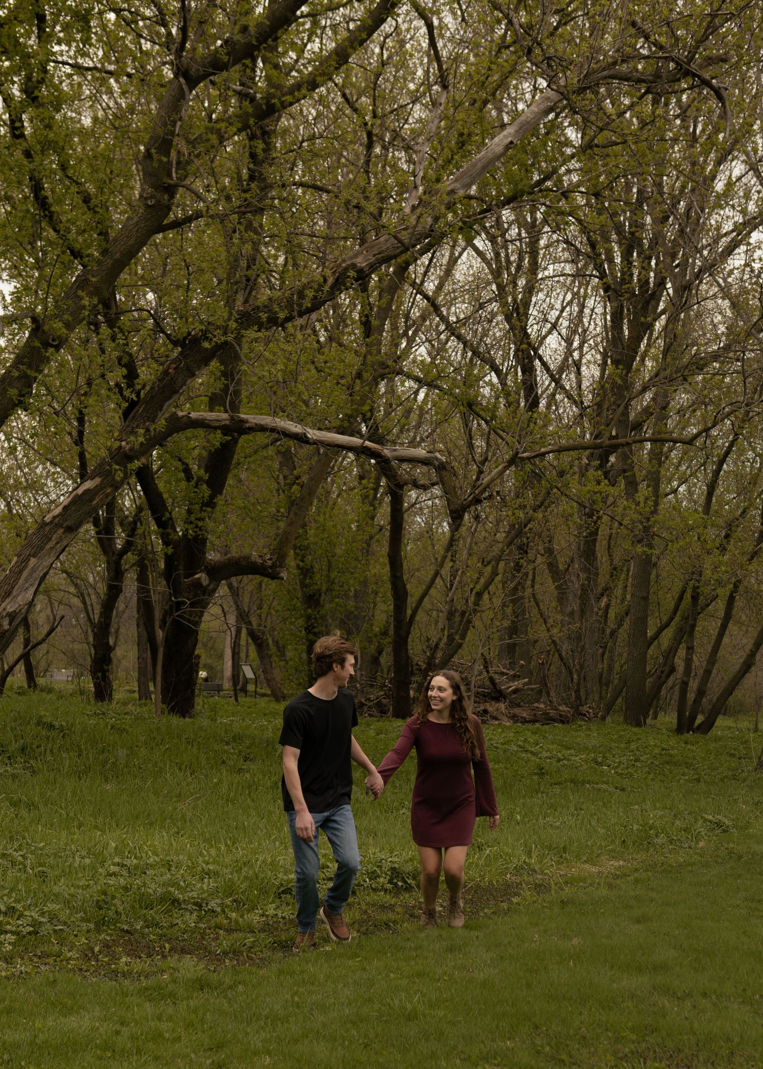 Couple holding hands and walking through a lush green forest clearing during spring engagement photo session.