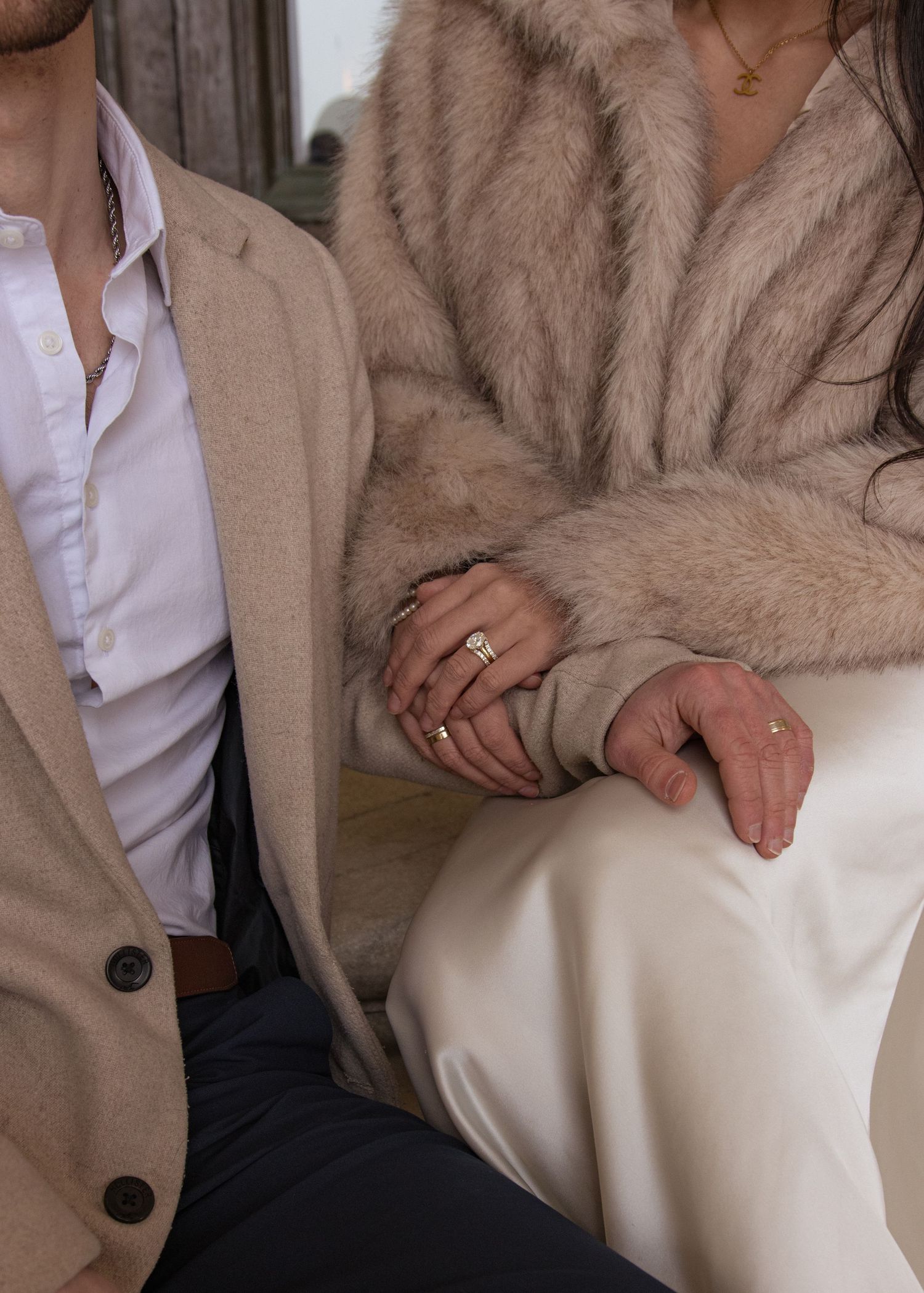 Couple holding hands, woman wearing fur coat and gold rings, man in beige blazer and white shirt, sitting together.
