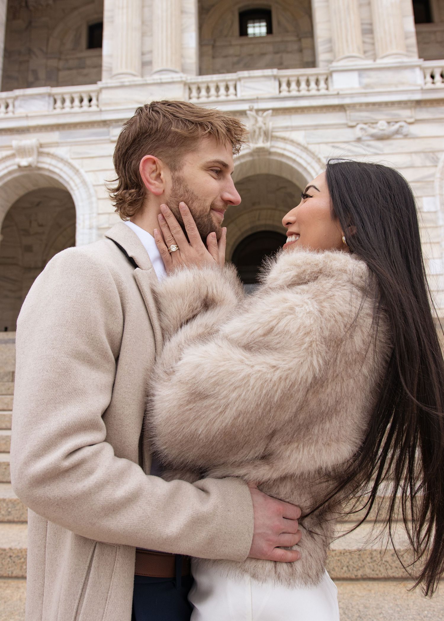 A romantic couple in light winter attire sharing tender moments in front of a grand stone building.