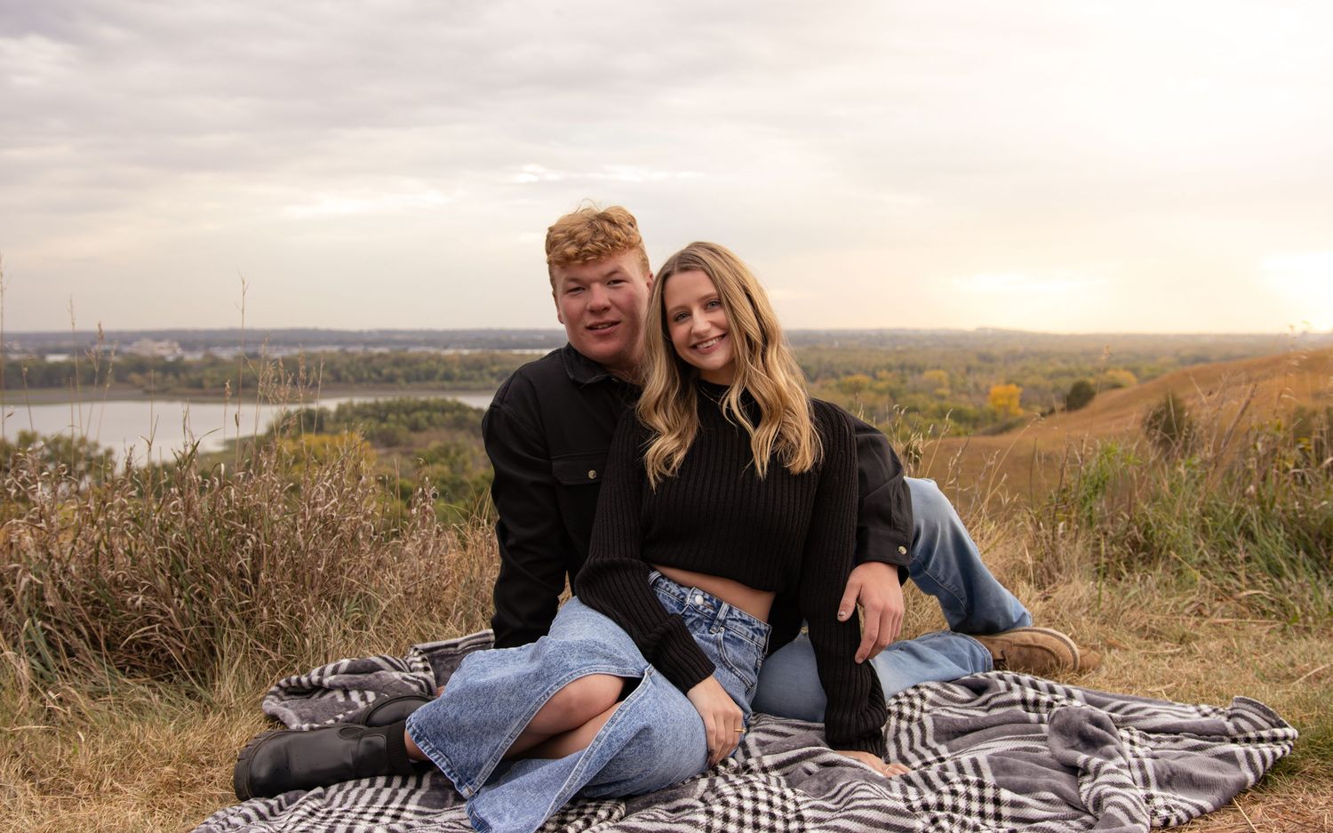 A couple relaxes on a blanket overlooking a scenic autumn landscape at sunset.