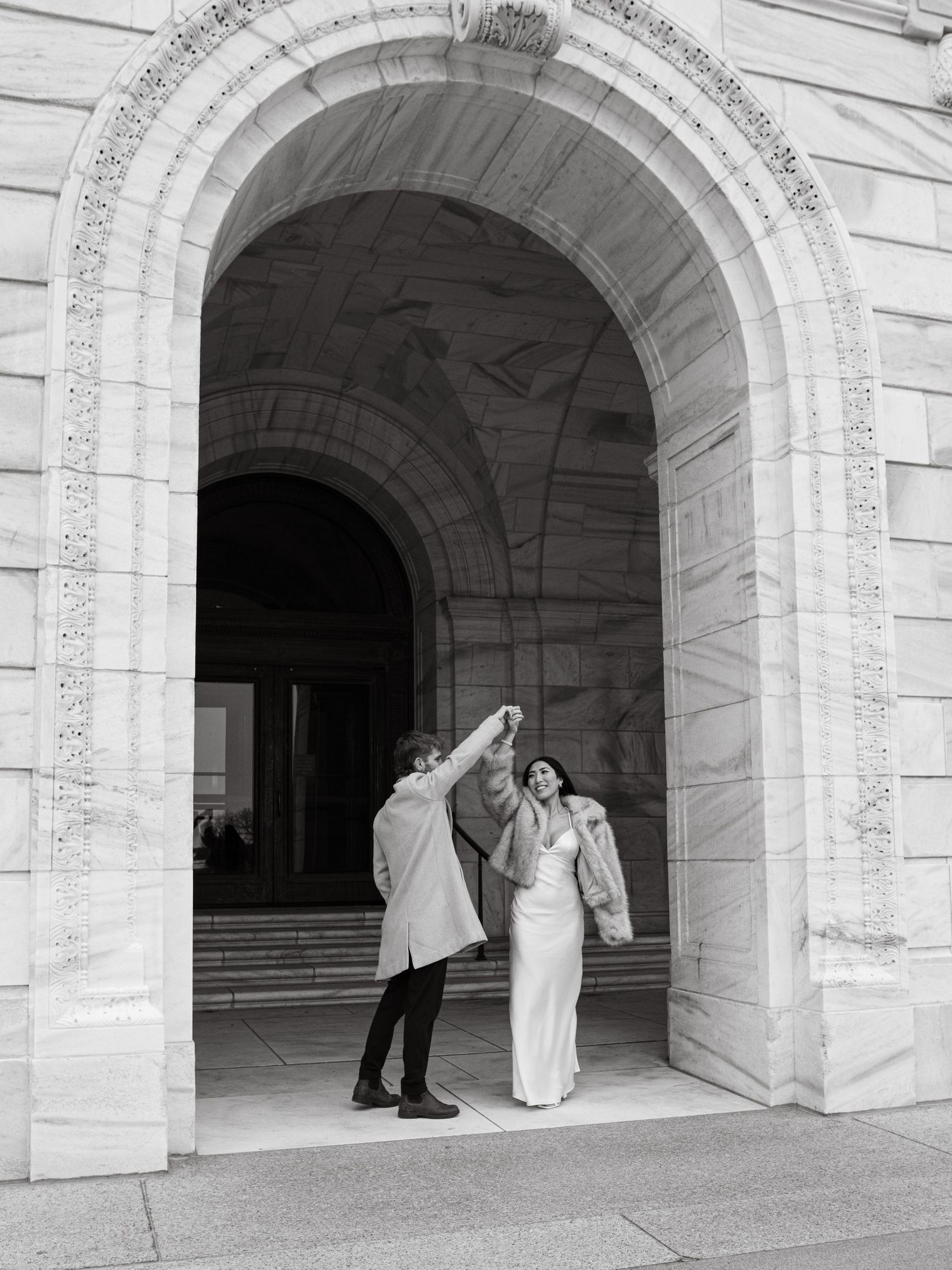 Black and white photo of a couple dancing under a grand stone archway, bride in white gown, groom in long coat.