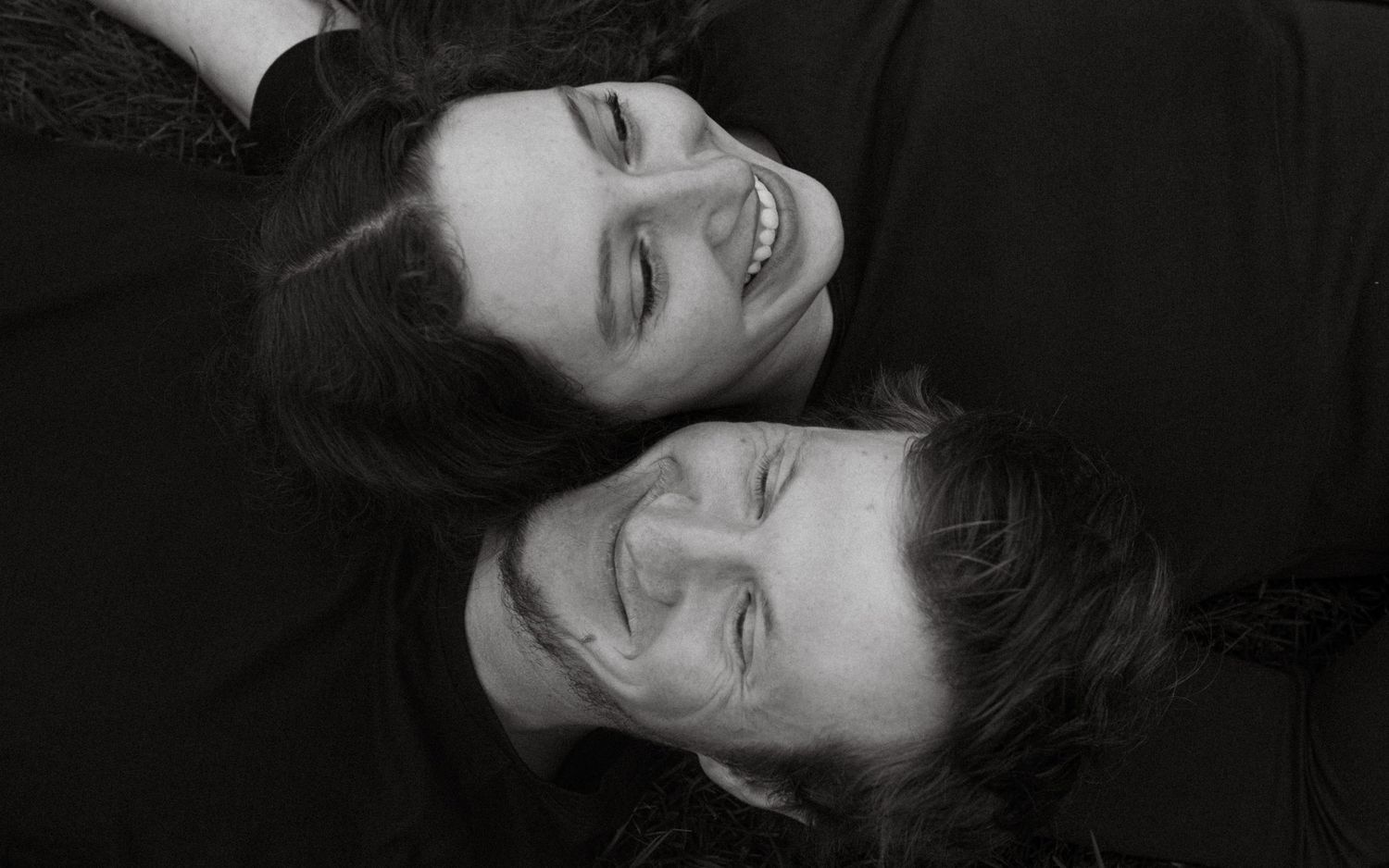 Black and white photo of two smiling people lying down with heads together, sharing a joyful moment.
