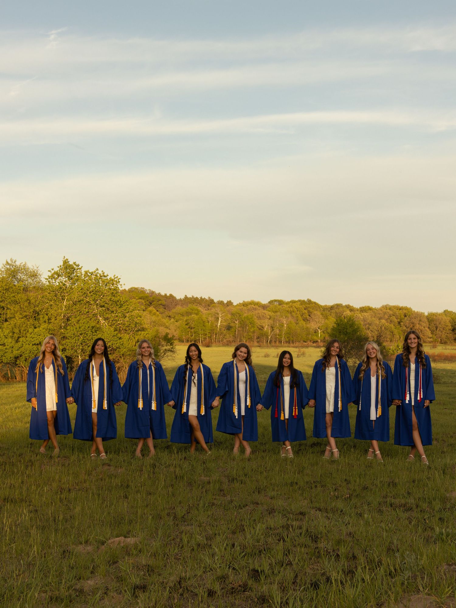 Ten female graduates in blue caps and gowns pose in a line outdoors in a sunny green field.