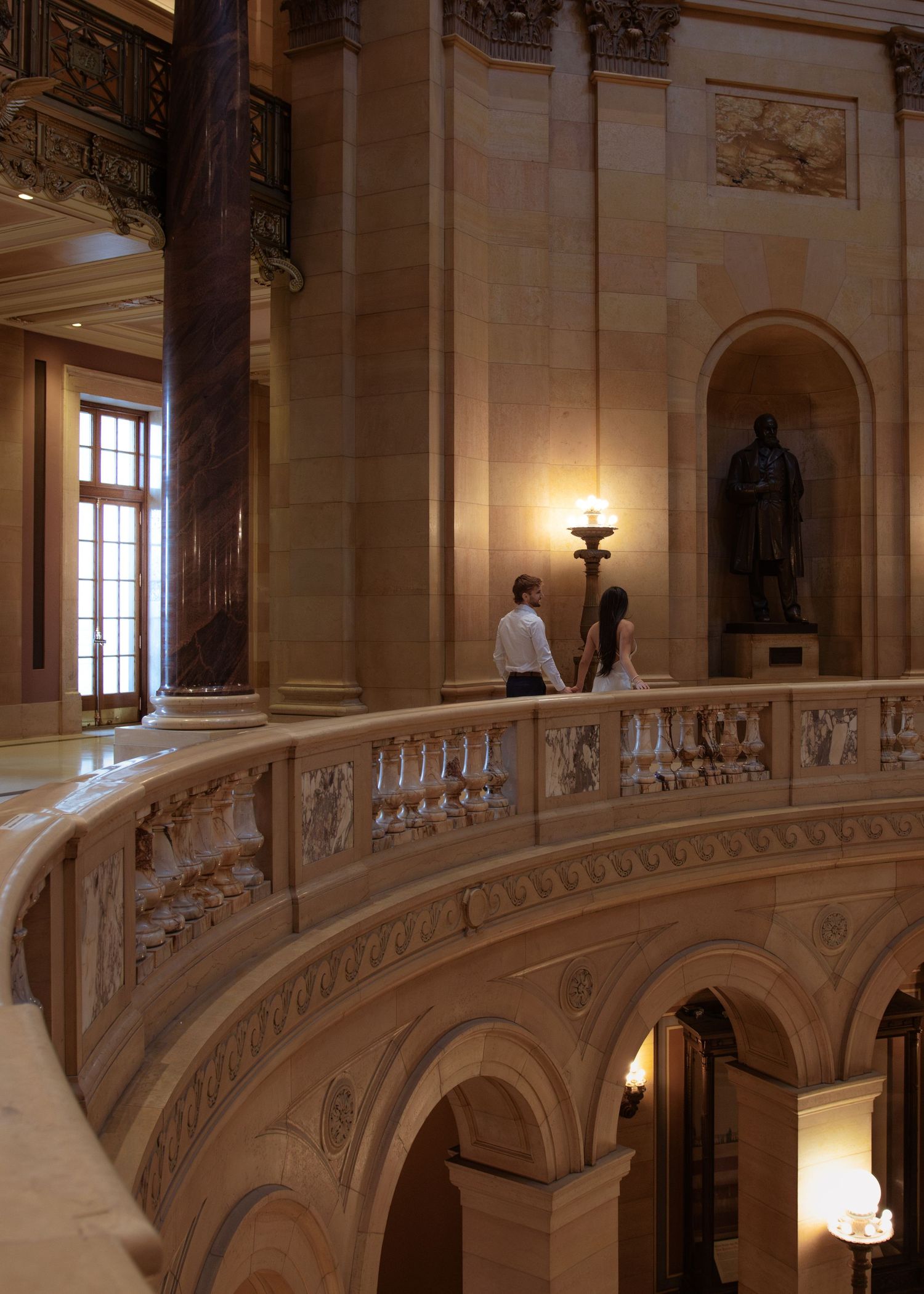Couple holds hands on ornate curved balcony inside grand neoclassical building with marble columns and statue.