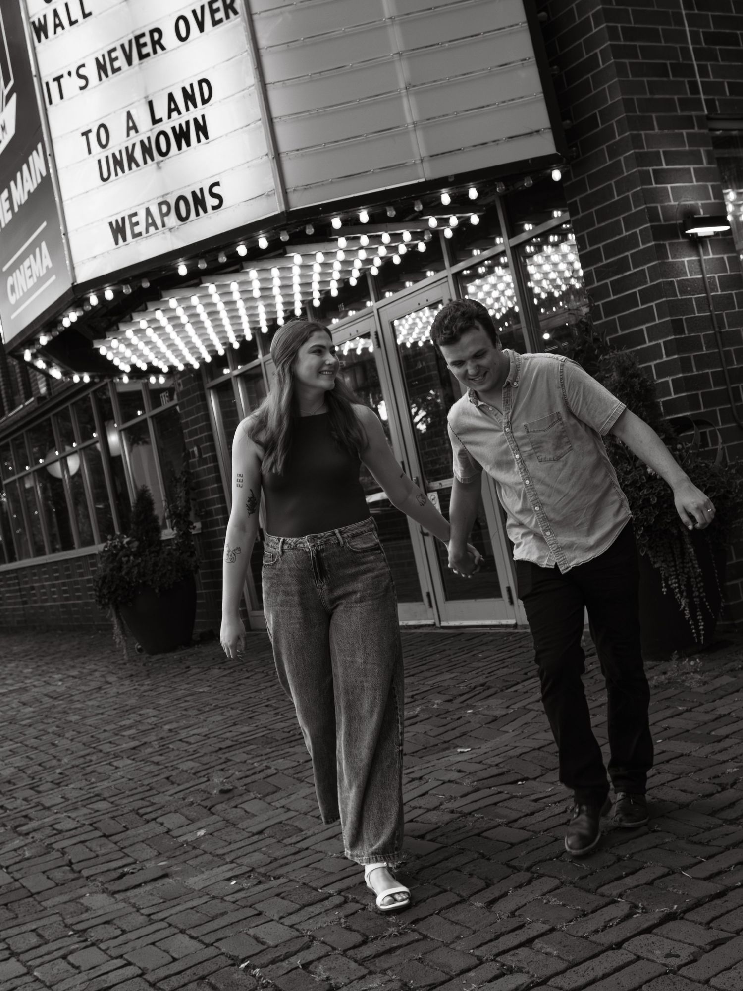 Black and white photo of people walking past illuminated movie theater signs.