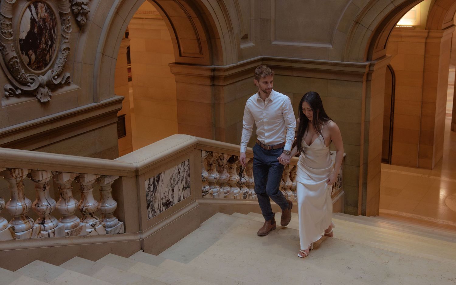A couple descends an elegant marble staircase in a grand historic building with ornate architectural details.