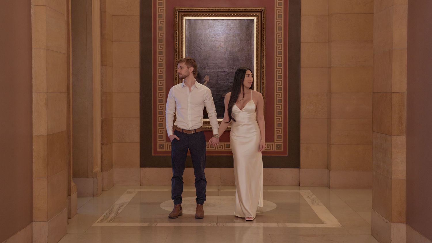 Elegant couple holding hands in a grand marble hallway, woman in white gown, man in casual attire near classic portrait.
