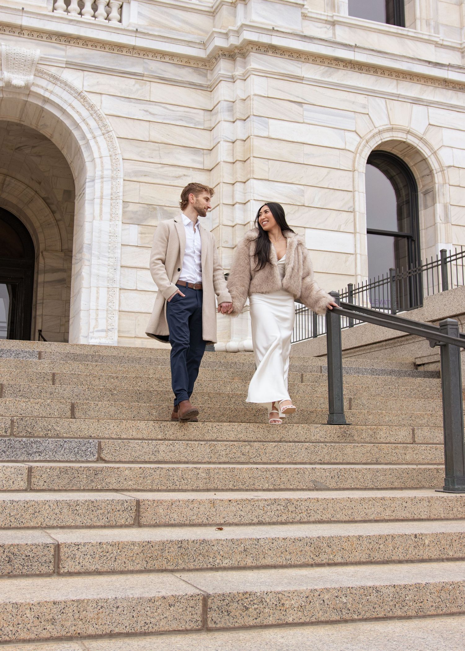 Couple holding hands walking down stone steps of elegant building, wearing neutral toned coats, white dress, and navy pants.