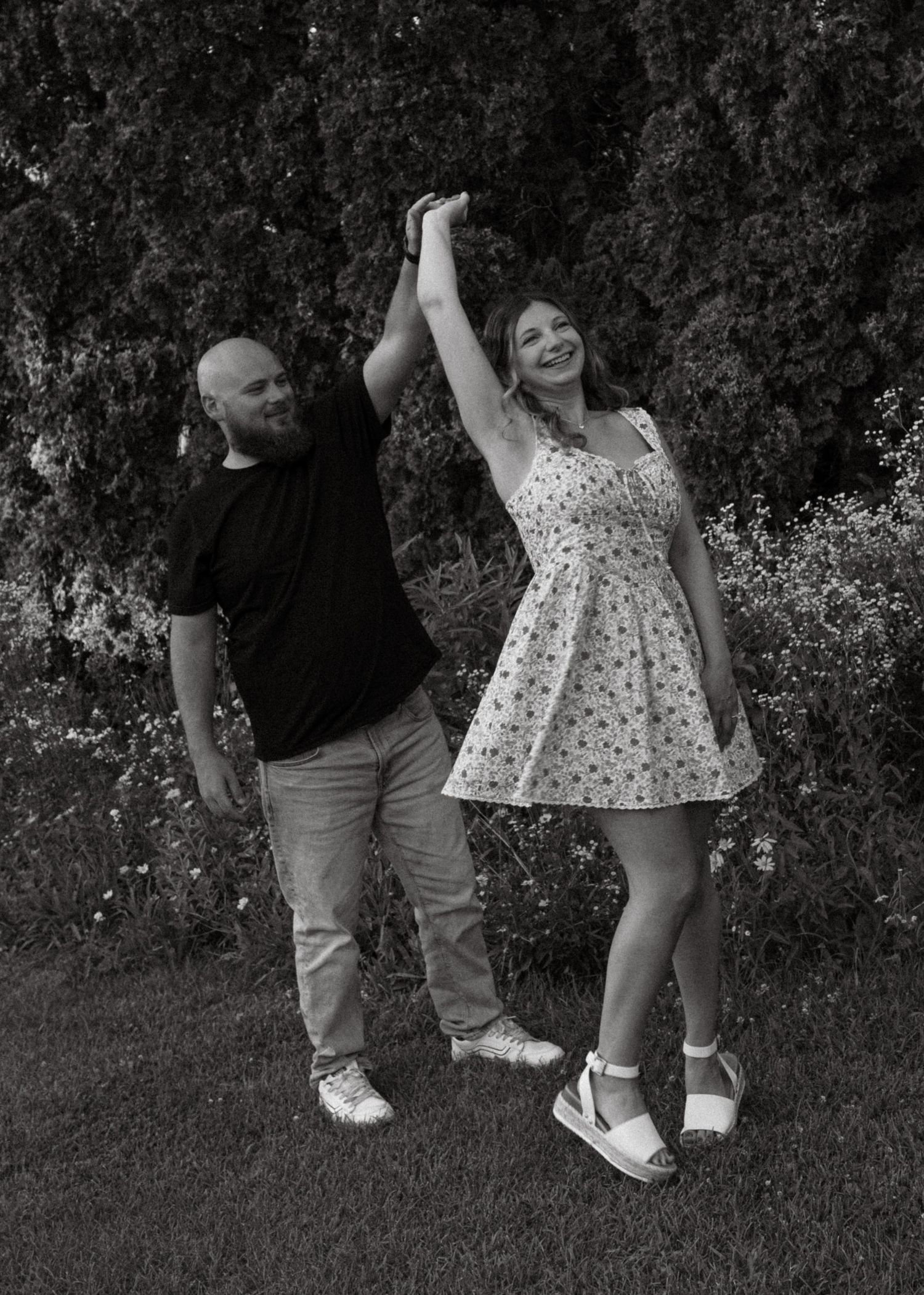 Black and white photo of a couple dancing outdoors, man spinning woman in floral dress near lush green trees.