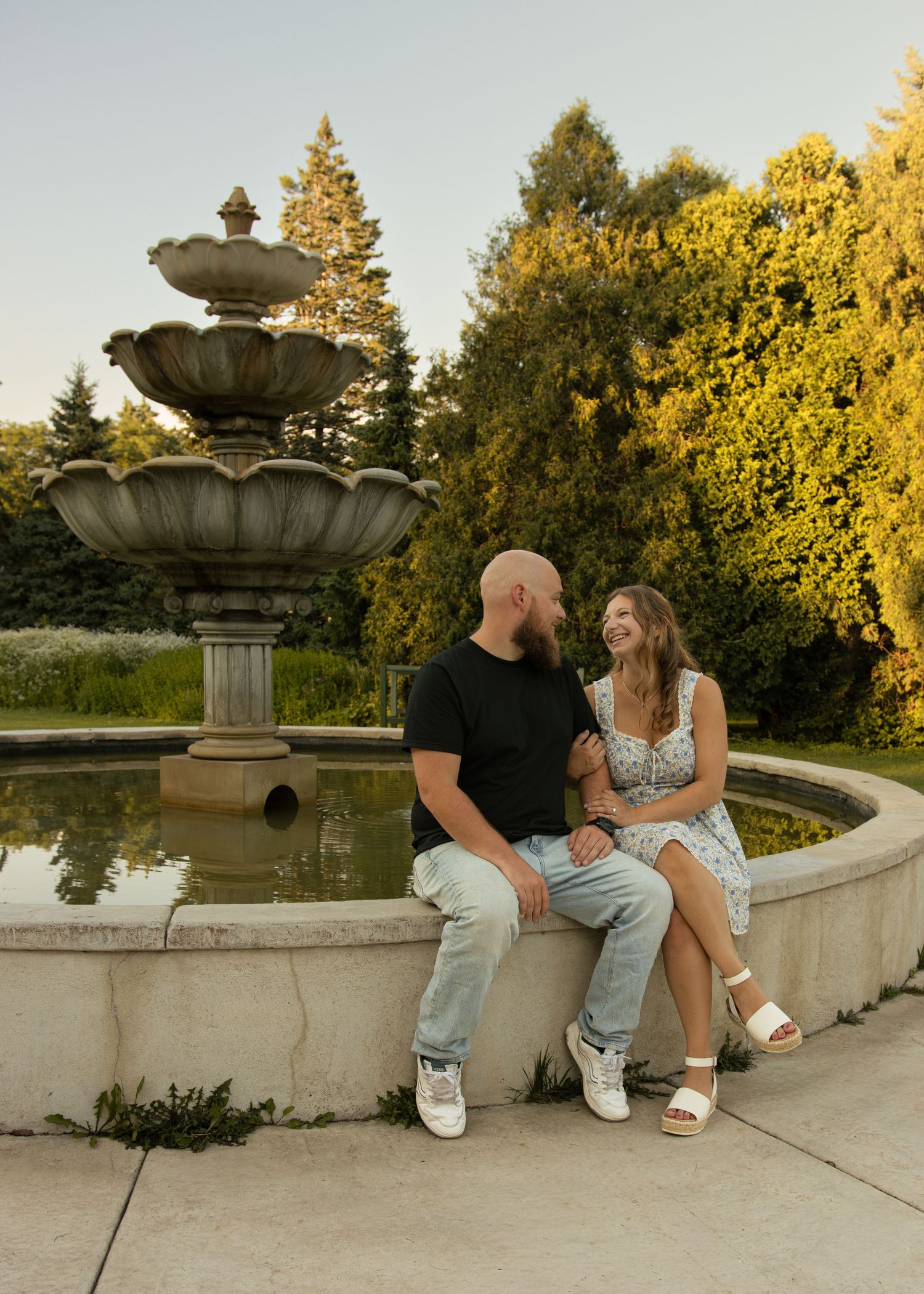 Couple sitting together by a stone fountain in a lush garden, smiling at each other in warm golden hour light.