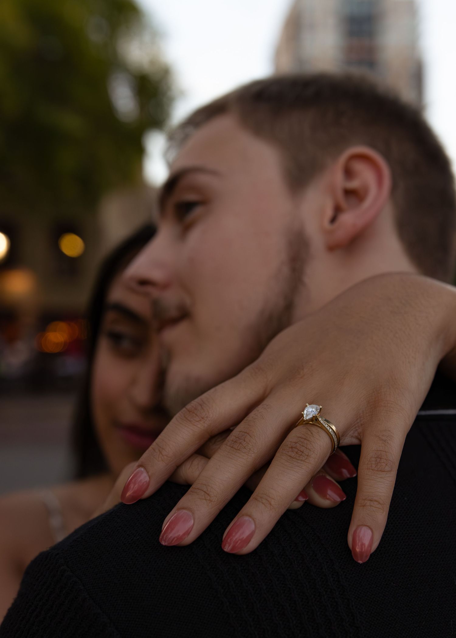 Couple embracing outdoors, woman displaying diamond engagement ring with pink nails on man's shoulder at dusk.