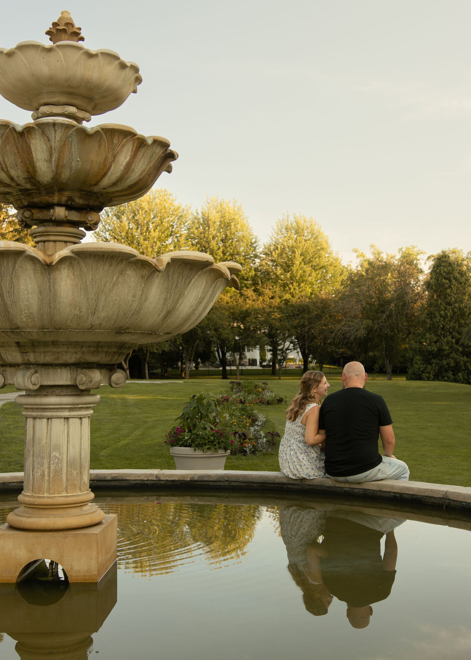 Couple sitting together by an ornate stone tiered fountain in a lush green park at golden hour.
