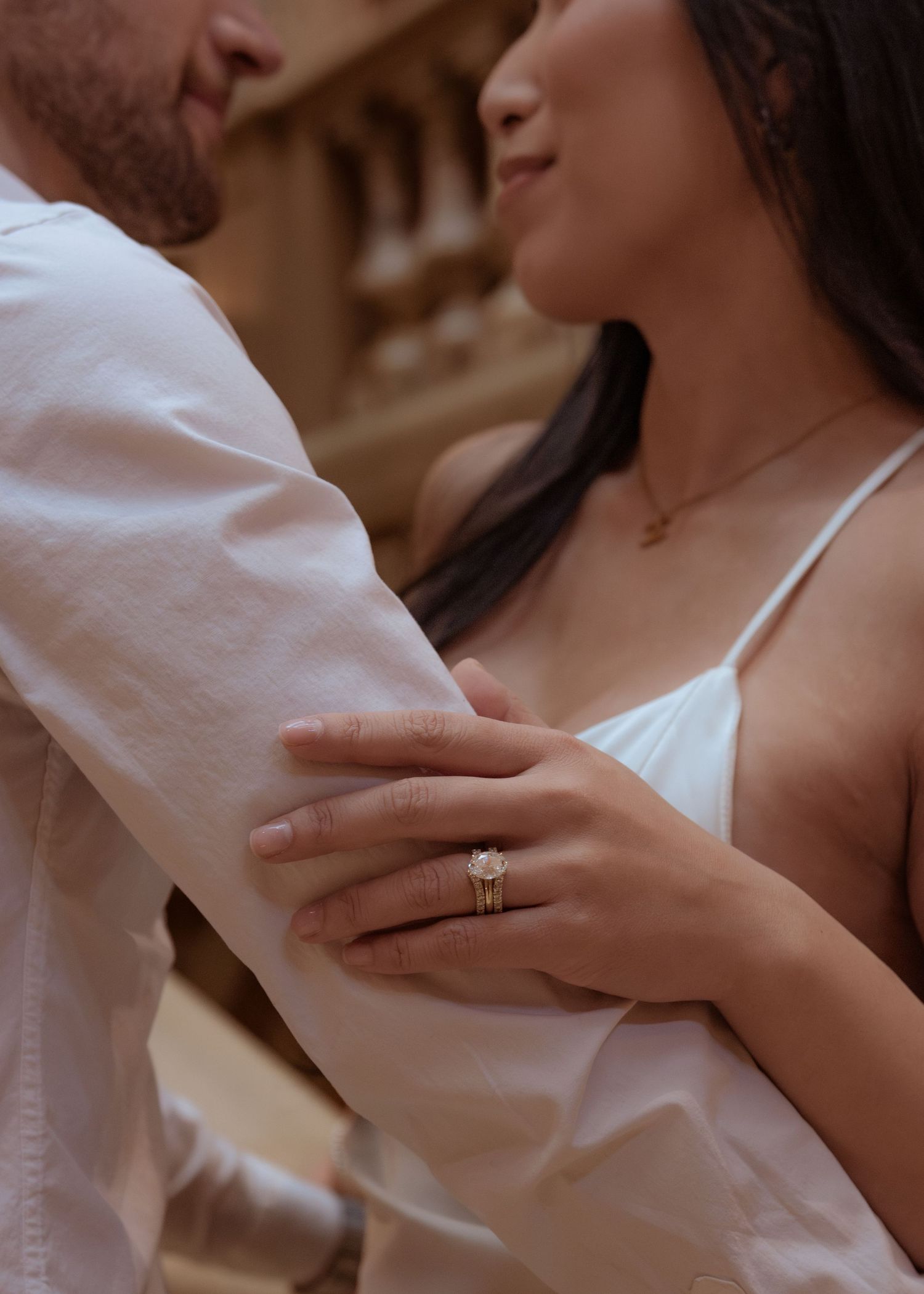 Couple embracing outdoors, woman wearing a diamond engagement ring and gold necklace, dressed in white.