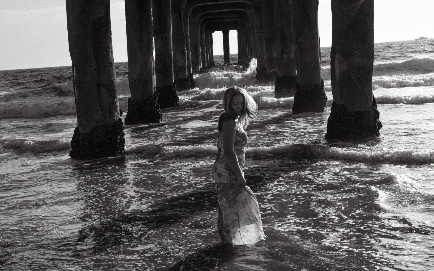 Black and white photo of a woman in a white dress standing in ocean waves beneath a wooden pier.