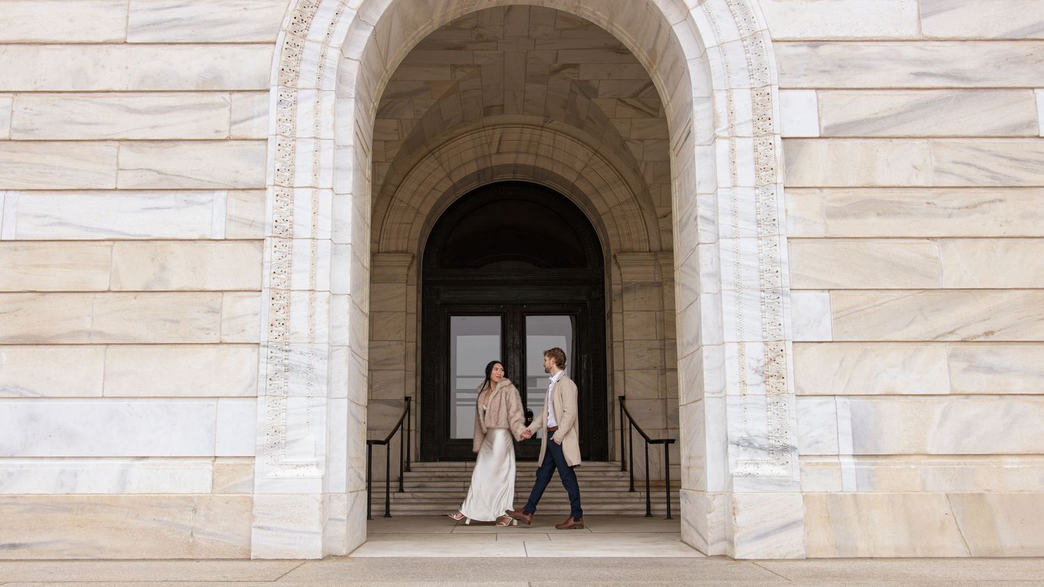 Couple walks hand in hand through grand stone archway of historic marble building on wedding day.