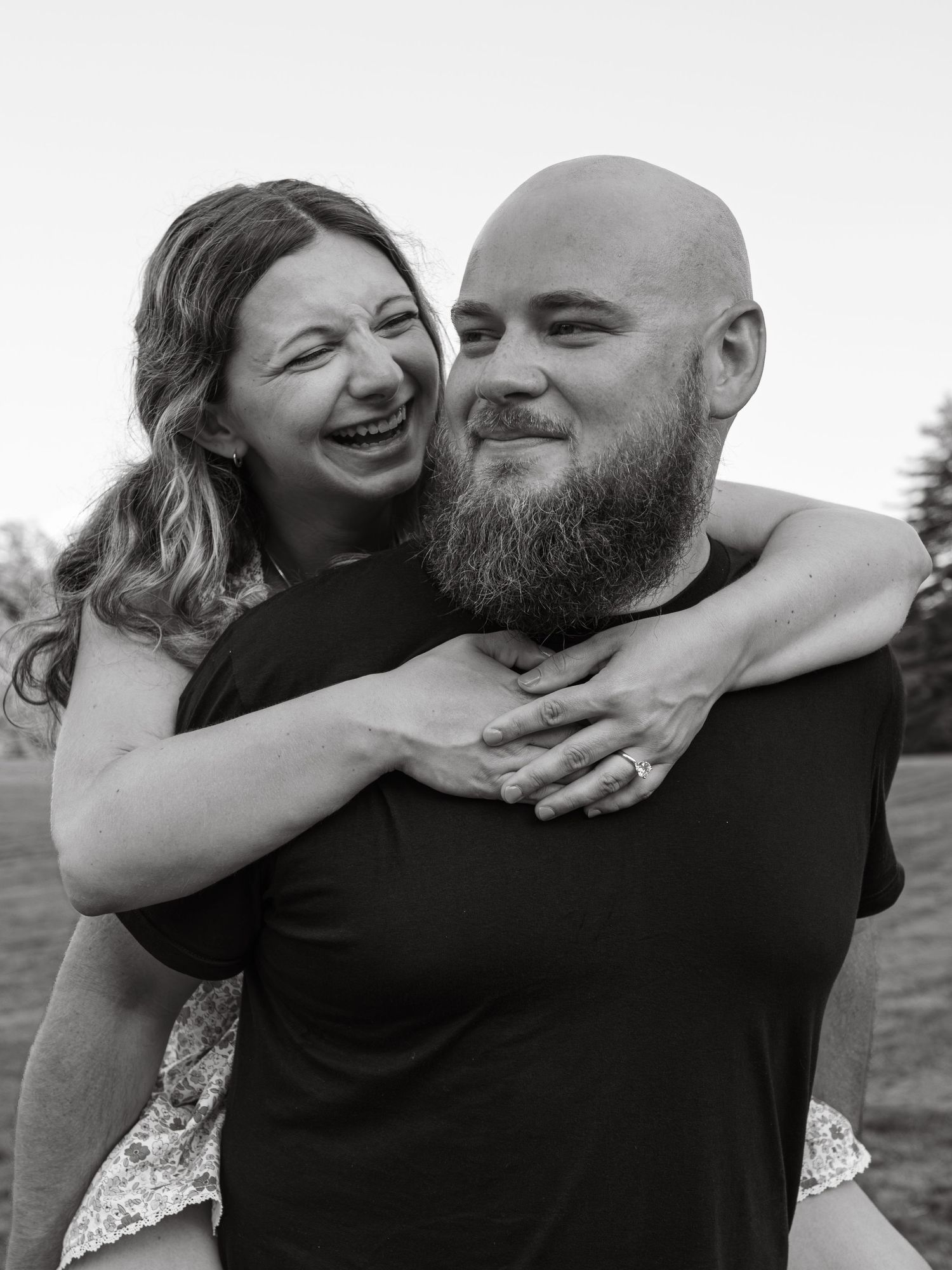 Black and white photo of a laughing woman hugging a bald bearded man from behind outdoors.