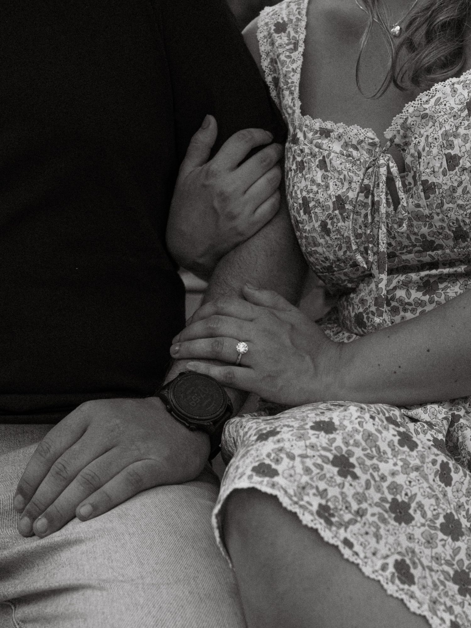 Black and white photo of a couple sitting together, woman in floral dress showing engagement ring, man wearing a watch.