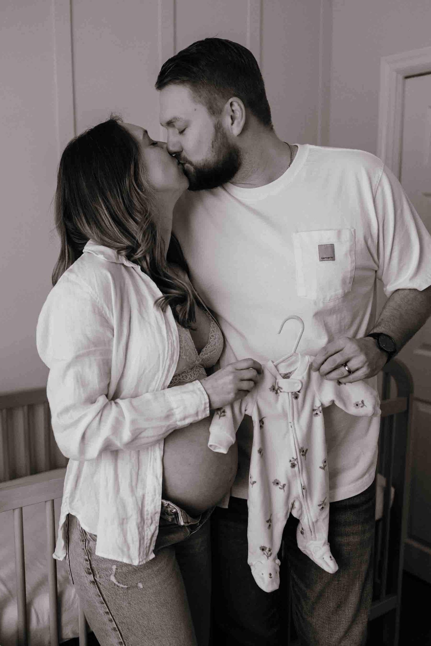 Expectant parents sharing a kiss while holding their baby's first outfit during an in-home maternity photography session in Toronto.