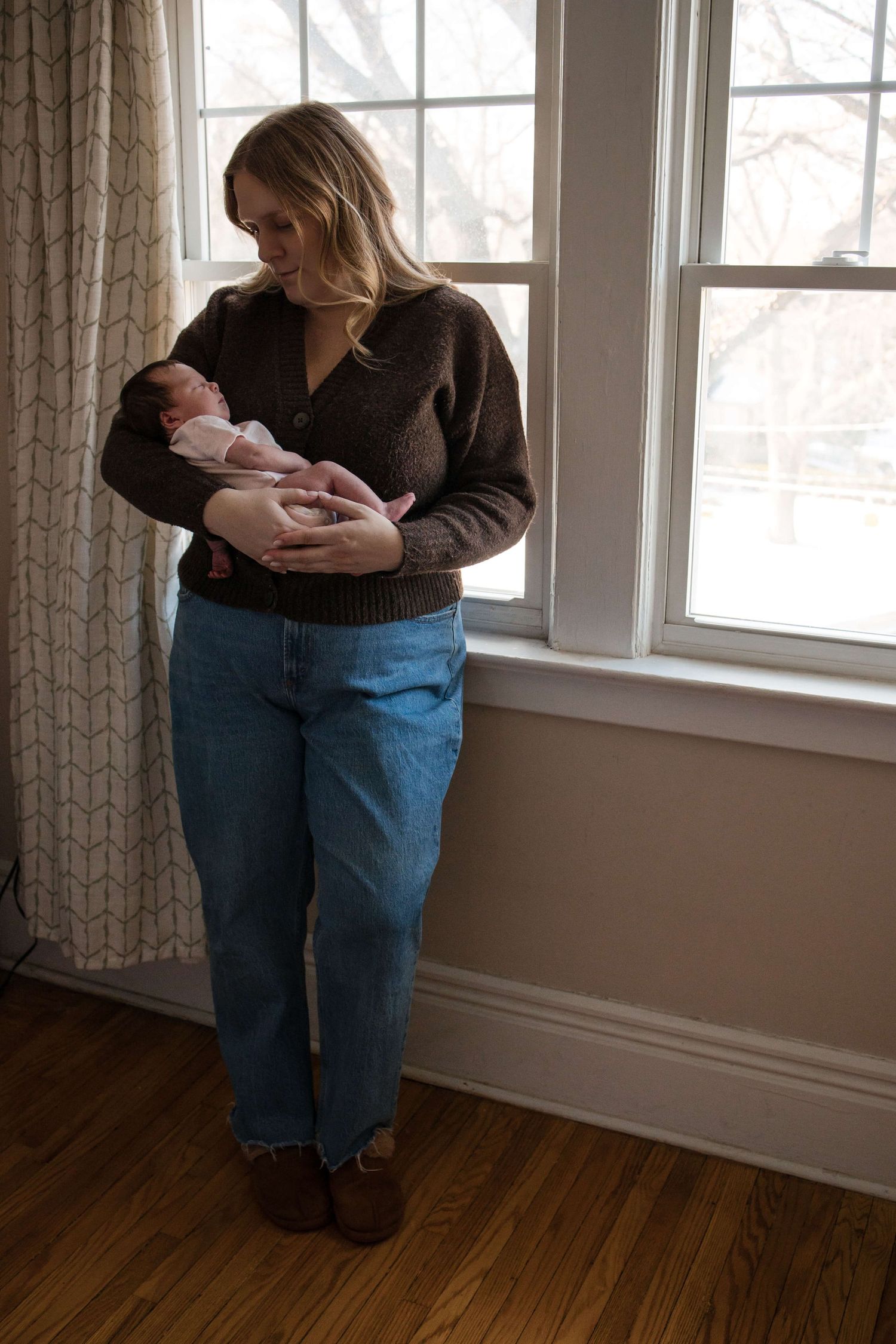 Mother holding her newborn by a window during an in-home newborn photography session in Toronto, capturing warmth, love, and connection.