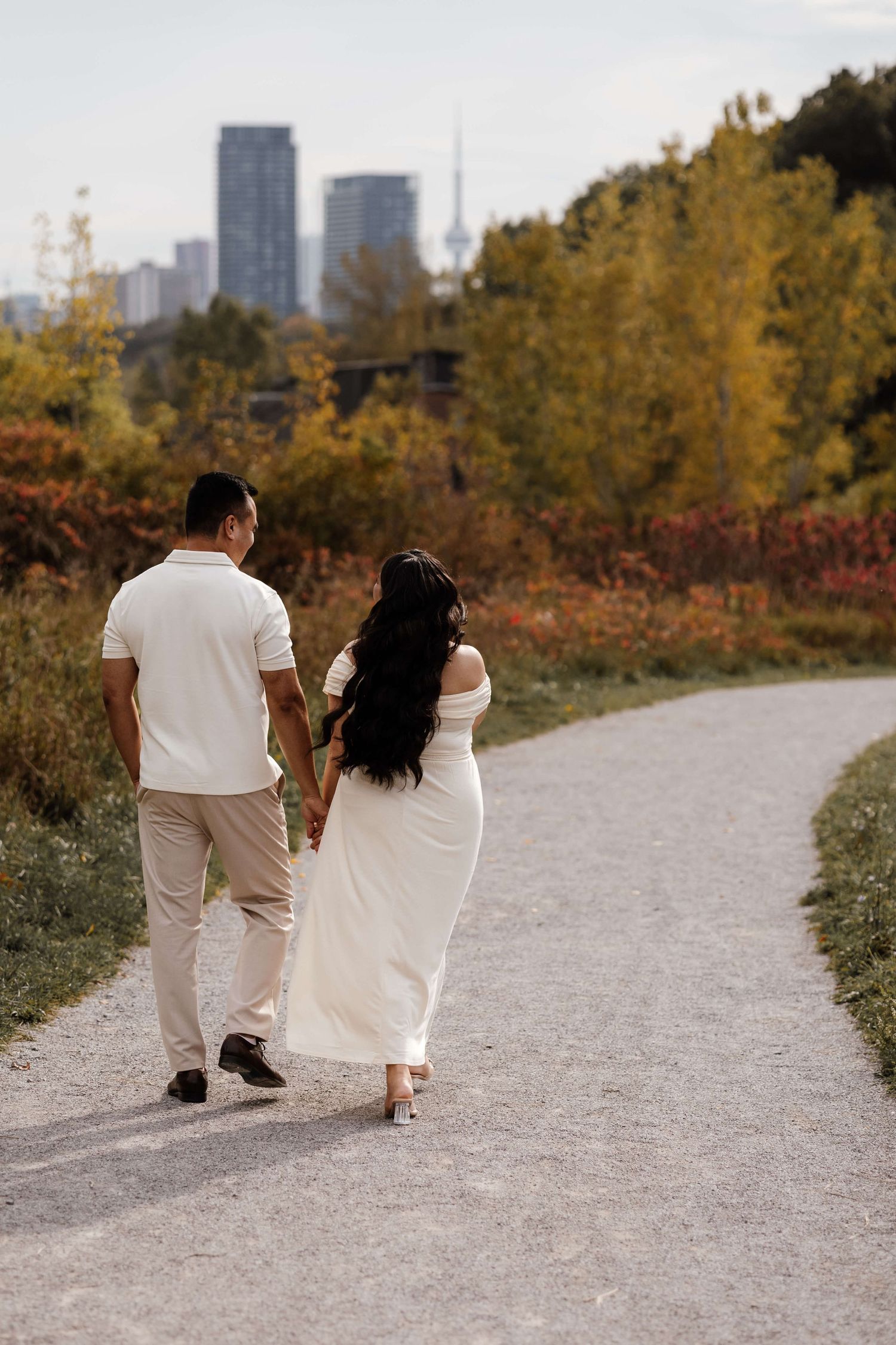 Expectant parents walking with the CN Tower in the background at Evergreen Brick Works during an outdoor maternity session in Toronto.