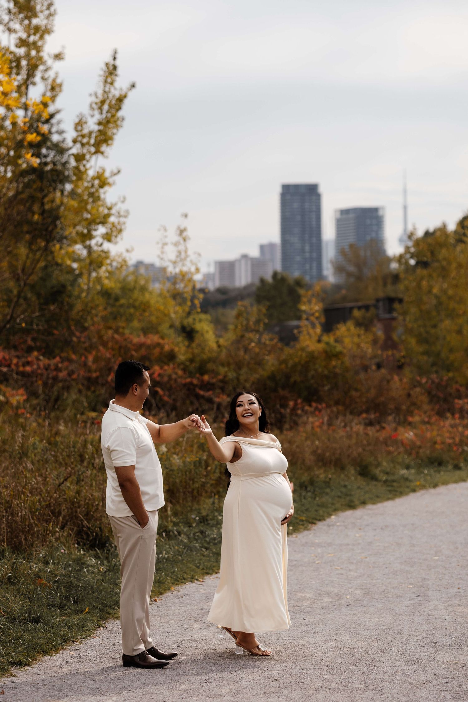 Expectant parents sharing a joyful moment at Evergreen Brick Works with the CN Tower in the background during maternity session in Toronto.