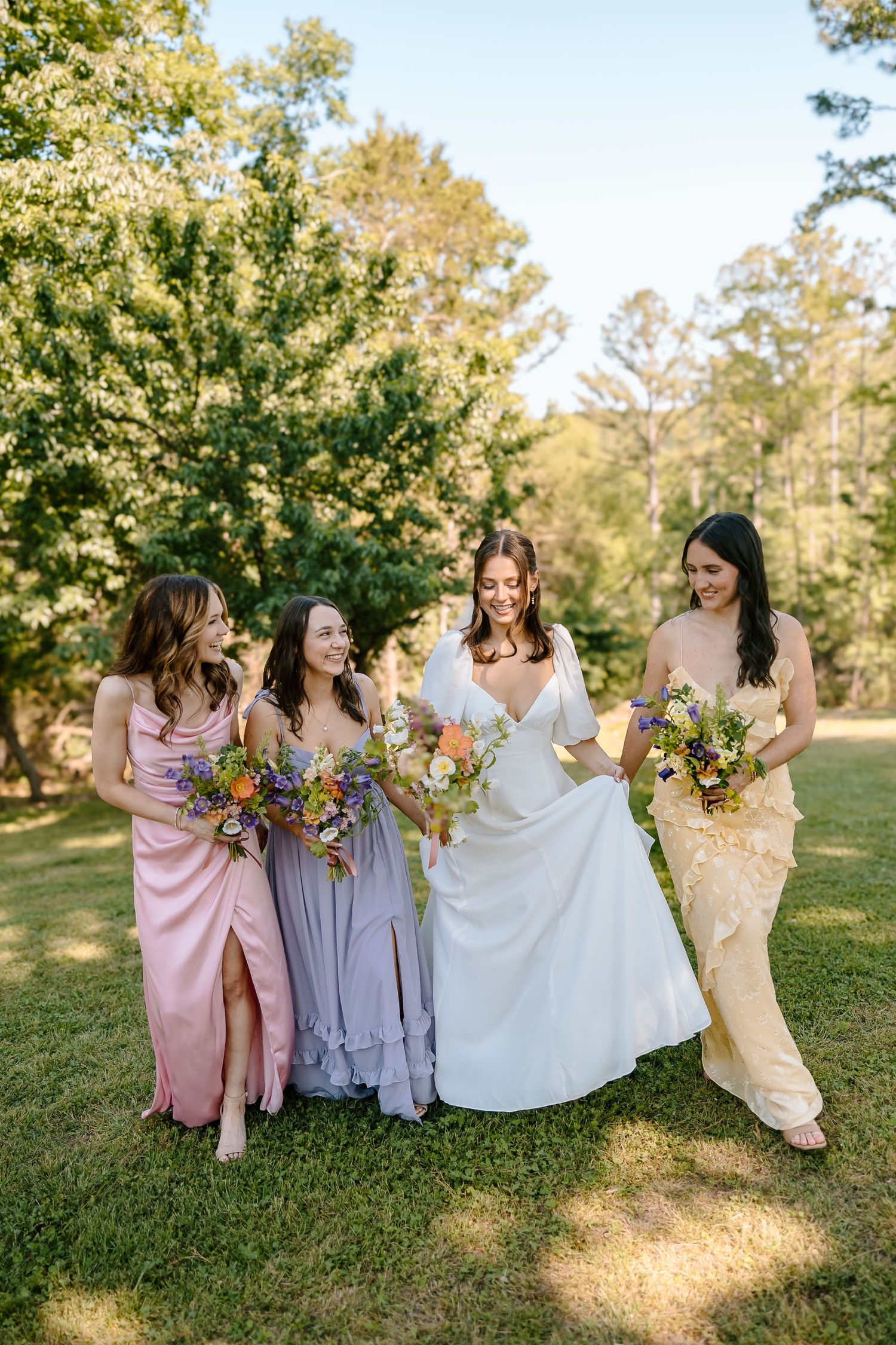 Bride in white gown with three bridesmaids in pink, lavender, and yellow dresses holding colorful bouquets outdoors.