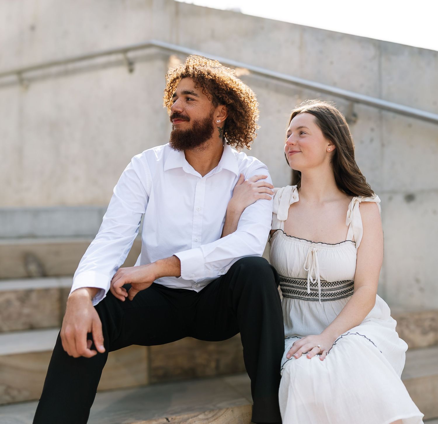 A smiling couple sits on outdoor steps, man in white shirt and woman in elegant white dress, enjoying a sunny day together.
