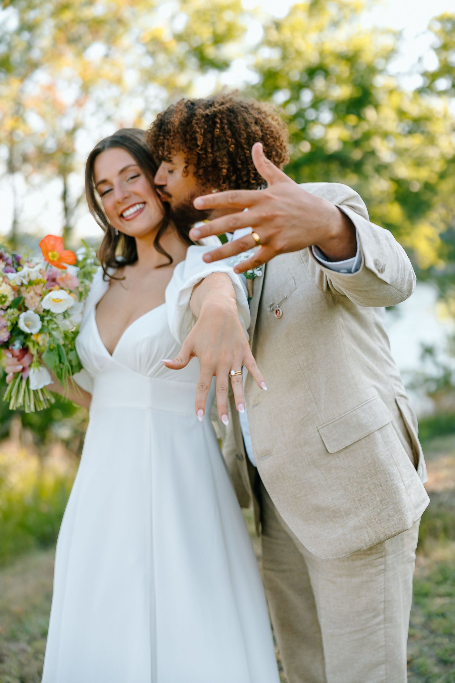Happy bride in white dress holds flowers as groom in tan suit embraces her and shows off wedding rings outdoors.