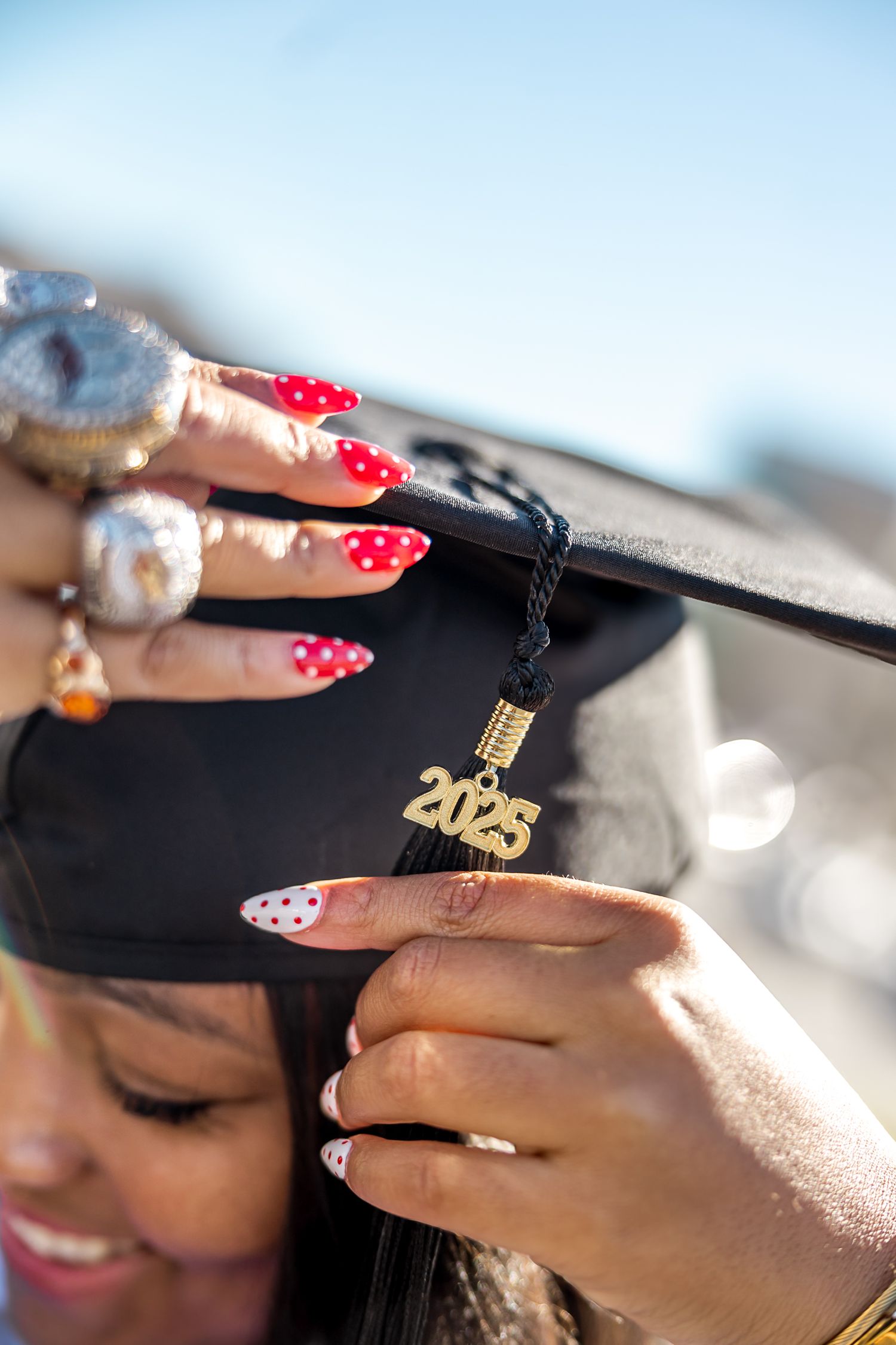 Close-up of hands adjusting graduation cap tassel with red polka dot nails and class of 2025 charm. philadelphia