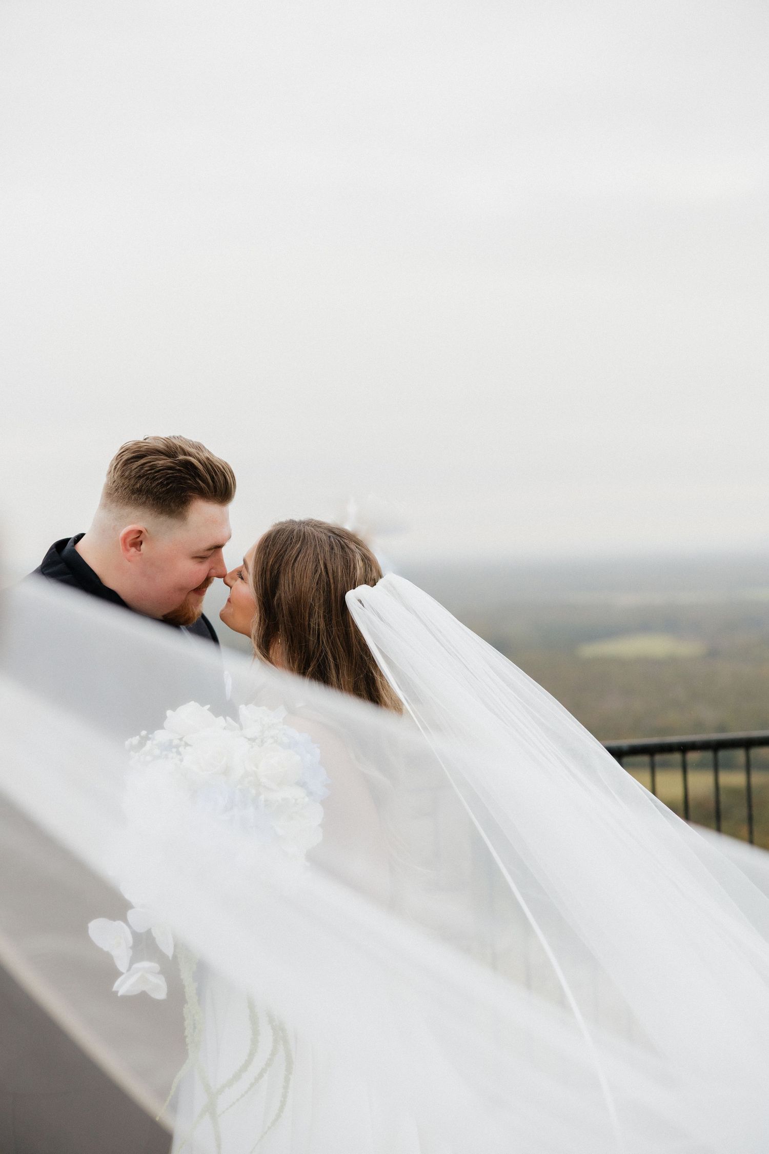 Couple embracing in intimate moment with flowing veil against cloudy sky.
