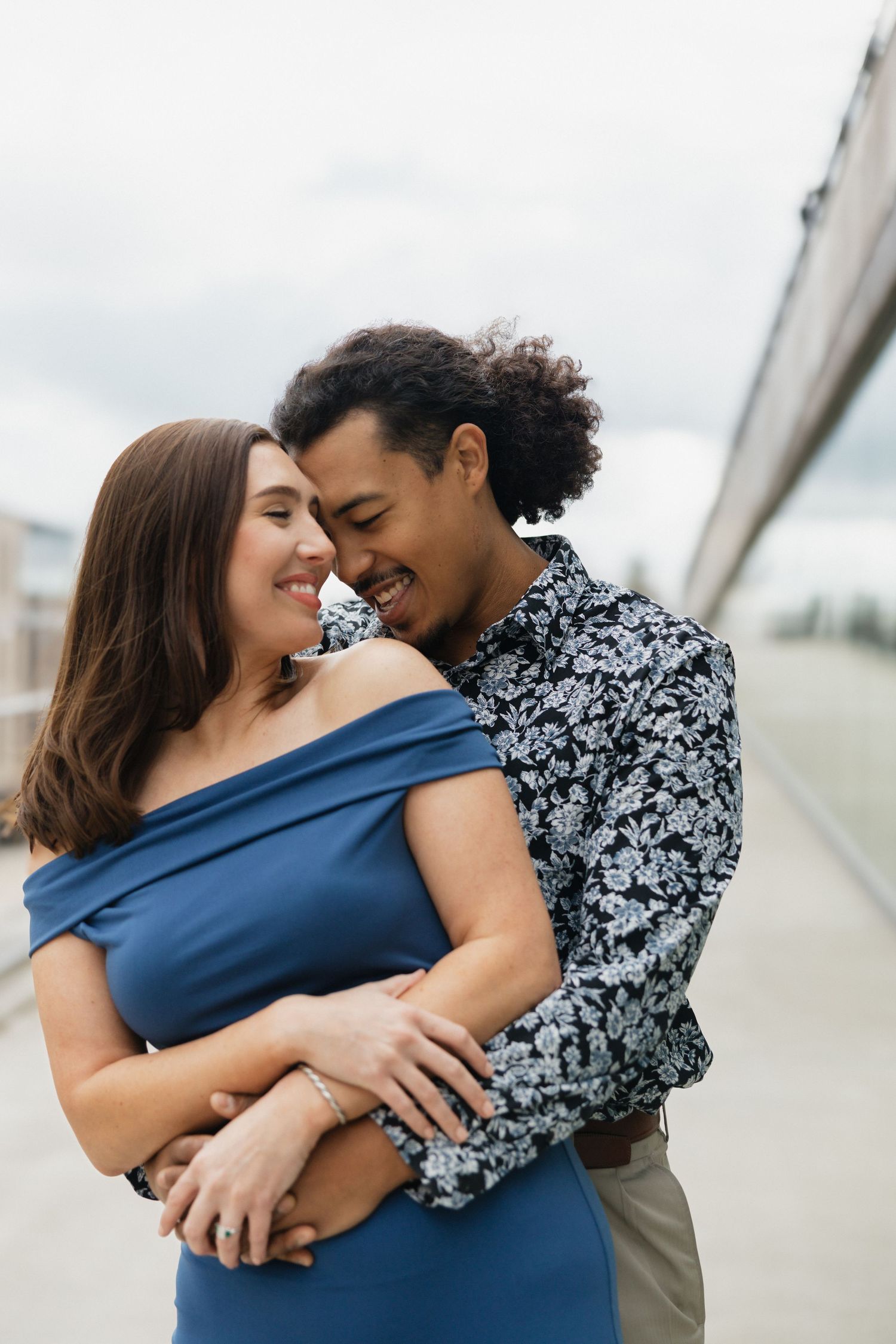 A happy interracial couple embracing outdoors, woman in blue off-shoulder dress, man in floral shirt, smiling together.