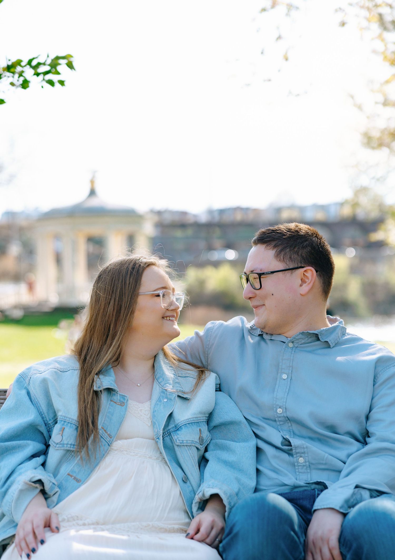 A young couple in denim jackets sit together in a sunny park, smiling at each other with a gazebo in the background.