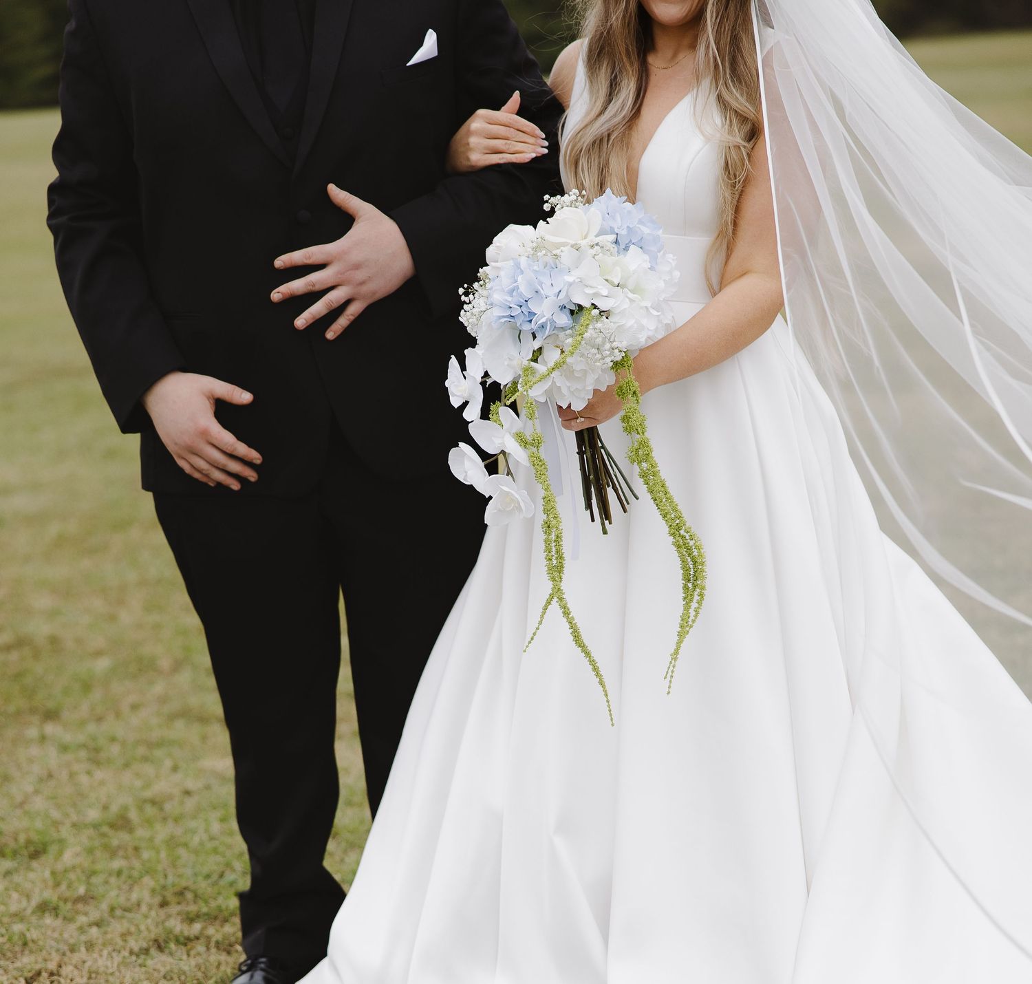 Bride in white ball gown holding blue hydrangea bouquet stands with groom in black suit on green lawn.