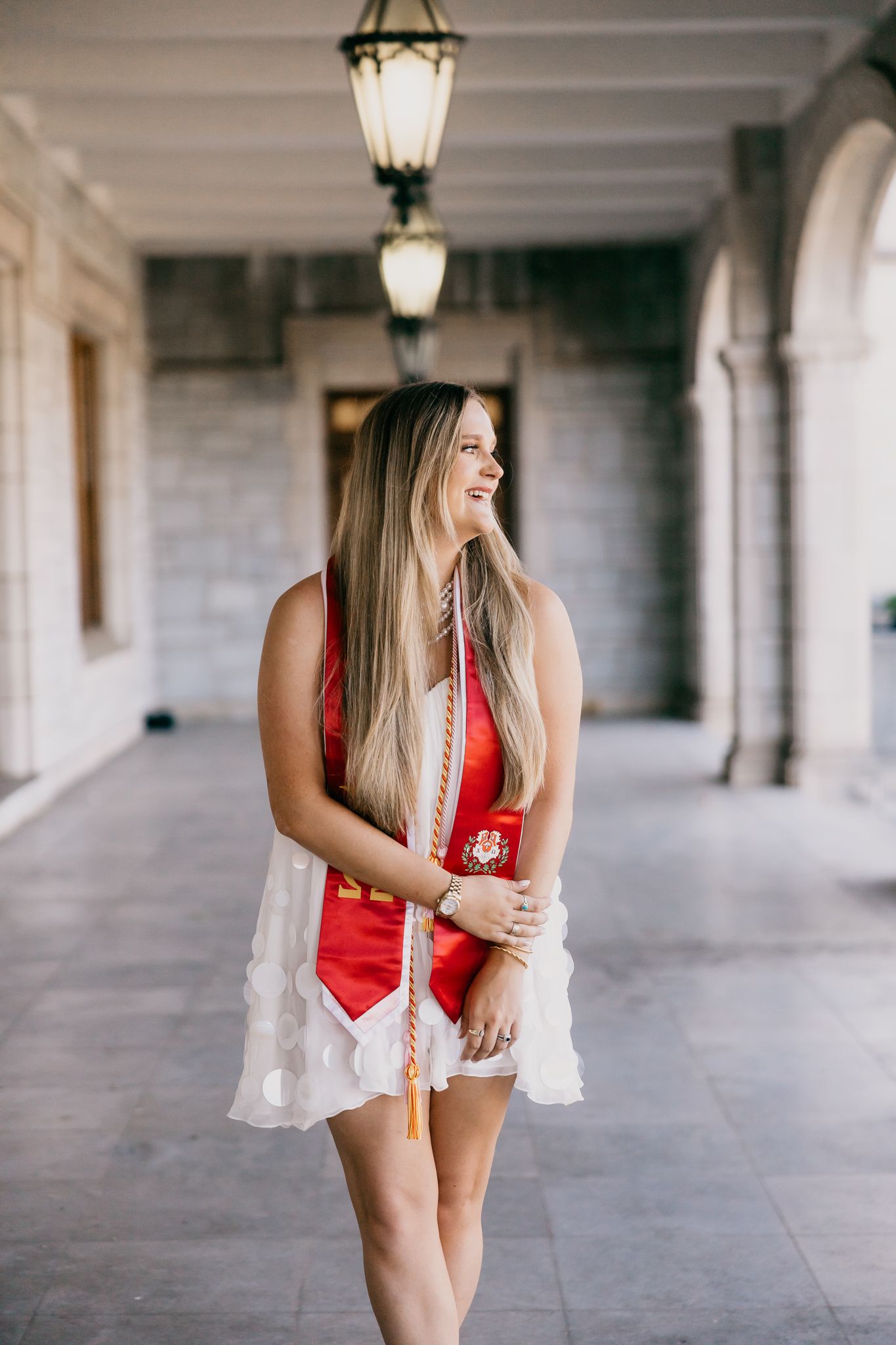 A person in a red outfit and white lace shawl poses in a corridor with arched columns and hanging lanterns.